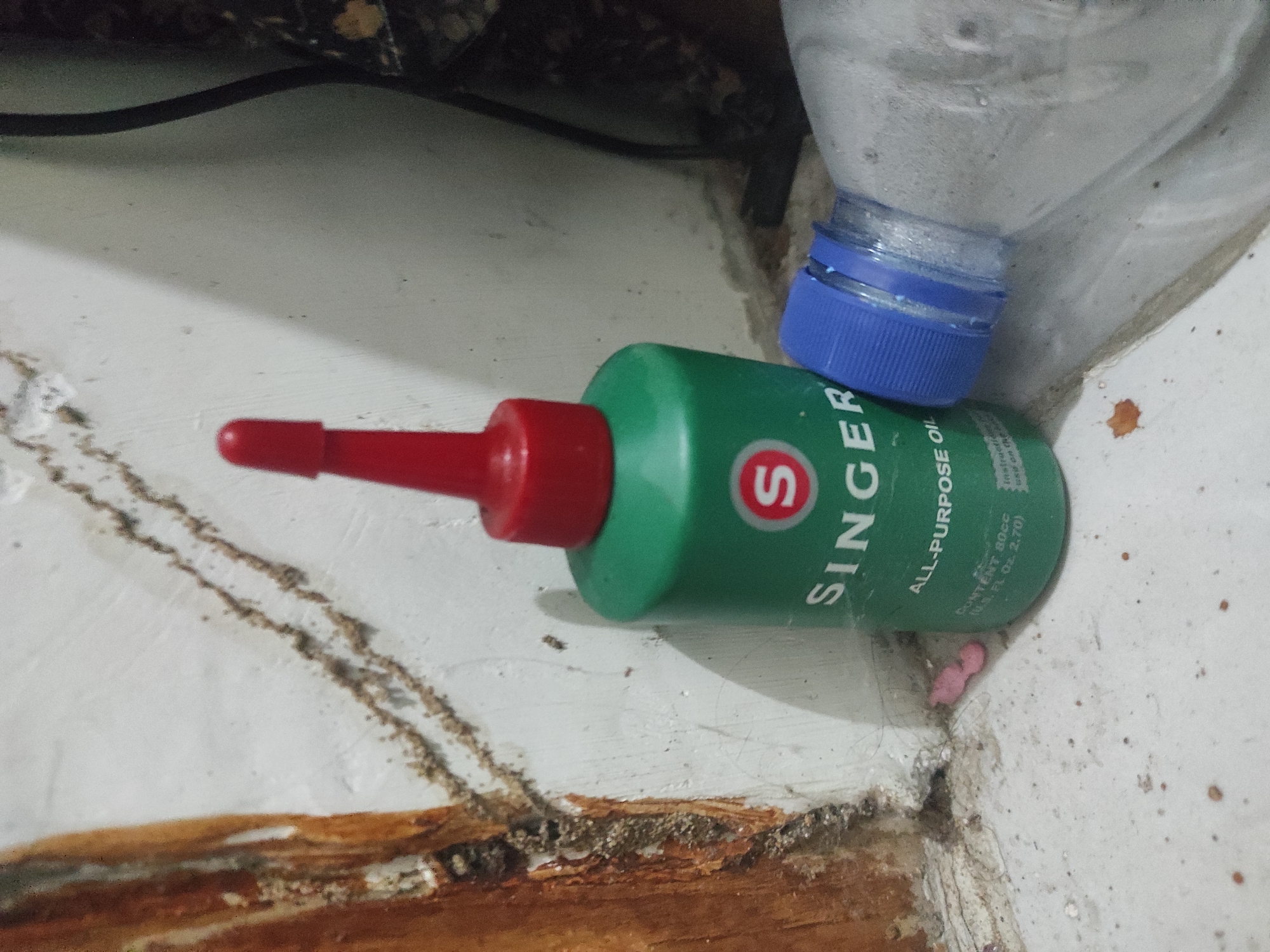 The image depicts a close-up, low-angle shot of a green Singer all-purpose oil bottle with a red cap, positioned on a white, dirty floor. The bottle is partially obscured by a clear plastic bottle with a blue cap. The environment appears to be an interior space with a textured wall, possibly a corner, showing signs of wear and dirt. To the left of the oil bottle, there is a section of what appears to be a wooden door frame with visible damage and debris. In the foreground, the floor is dusty and scattered with small brown specks. A small, pink, amorphous object is visible on the floor near the base of the oil bottle. The lighting is dim, casting shadows and highlighting the textures of the surfaces. The scene appears to be located in Balenrejo, Indonesia, based on the provided information. There are no people visible in the image.