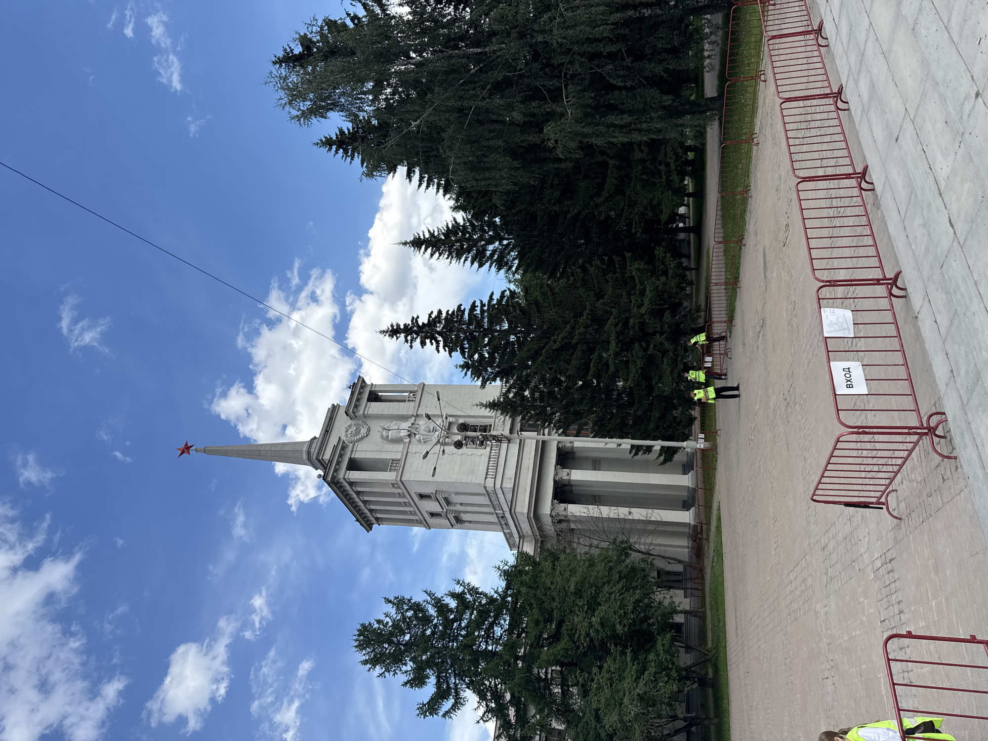 The image captures the Yekaterinburg Railway Station, a prominent landmark in the city. The station building, with its distinctive spire topped by a red star, is a grand, imposing structure constructed in a neoclassical style. Tall, manicured pine trees flank the building, contributing to the urban landscape. In the foreground, temporary red metal barriers are erected, delineating a pedestrian area. Several individuals, some wearing bright yellow safety vests, are visible near the barriers, suggesting some form of event or crowd management. The sky above is a vibrant blue, punctuated by fluffy white clouds, indicating fair weather. The overall scene depicts a public space within a city, with architectural significance and the presence of both natural elements and human activity.