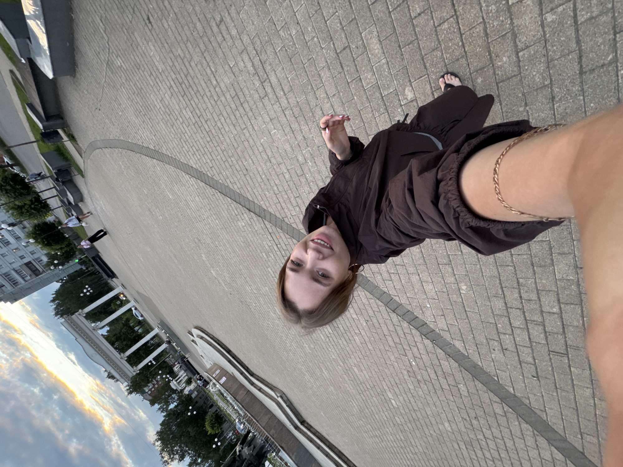 In Chaikovsky, Russia, a woman is taking a selfie in a paved public square. The sky above displays a dramatic sunset with colorful clouds. In the background, a large white archway with columns stands prominently, flanked by trees and buildings. Several other people are visible in the distance, some walking, others seated. A fountain is also present in the foreground left, with water spraying. The woman is wearing a dark brown jacket and dark pants, and she is smiling at the camera. Her right arm is extended, holding the phone for the selfie. A thin gold chain bracelet is visible on her left forearm.