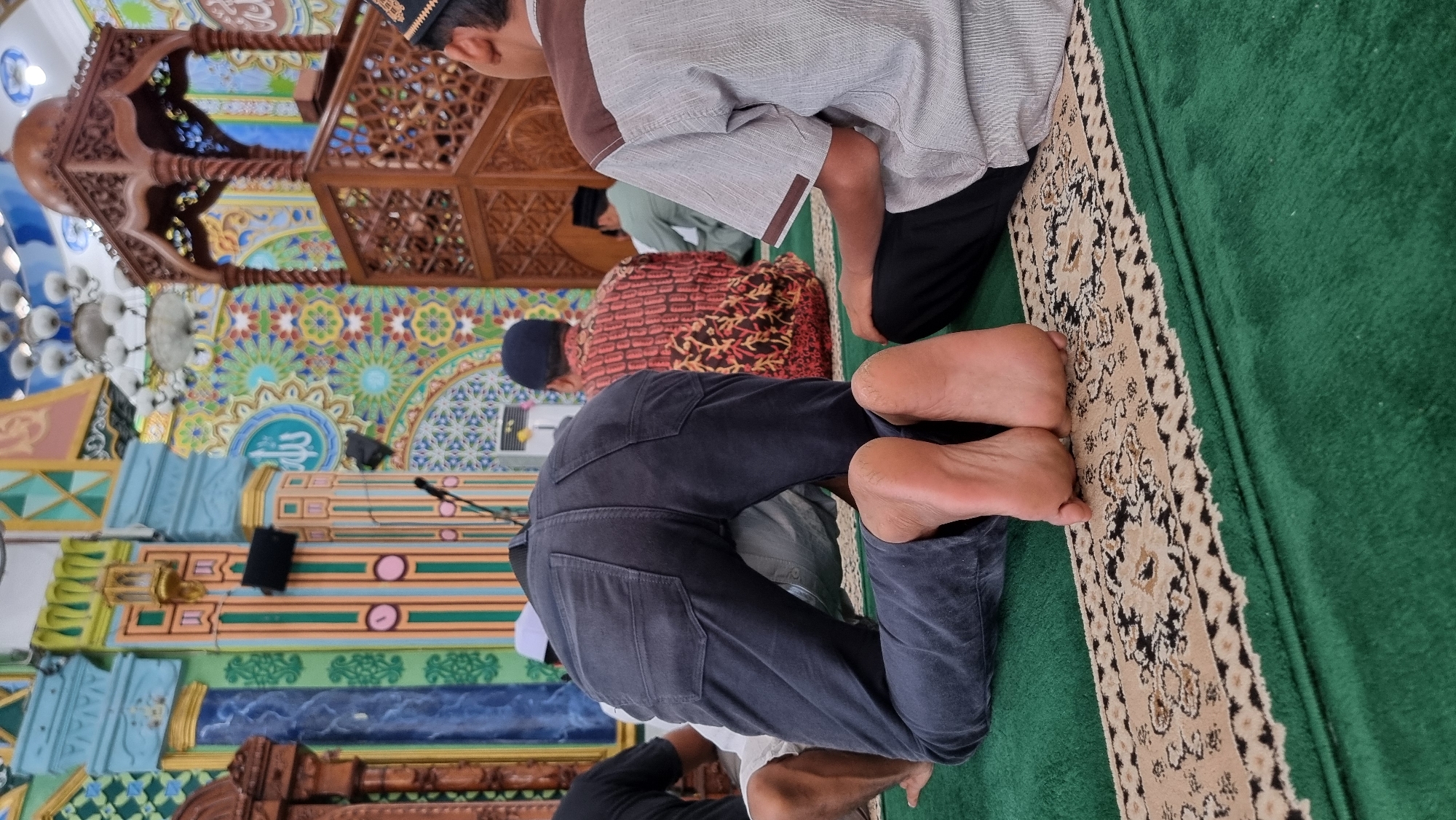 The image shows the interior of a mosque in Keudekruenggeukueh, Indonesia, during prayer. The focus is on the lower body of a person in the foreground, kneeling on a green carpet. This individual is wearing dark gray pants and their bare feet are positioned on a beige and black patterned rug. To the right, another person is seated, wearing a gray shirt with brown trim and black pants. In the background, several other individuals are also seated or kneeling in prayer. The mosque's interior features colorful, intricate wall decorations with geometric patterns and Arabic calligraphy. A wooden minbar, or pulpit, is visible on the right side of the image, and ornate chandeliers hang from the ceiling. The overall scene depicts a religious gathering during prayer time within a mosque.