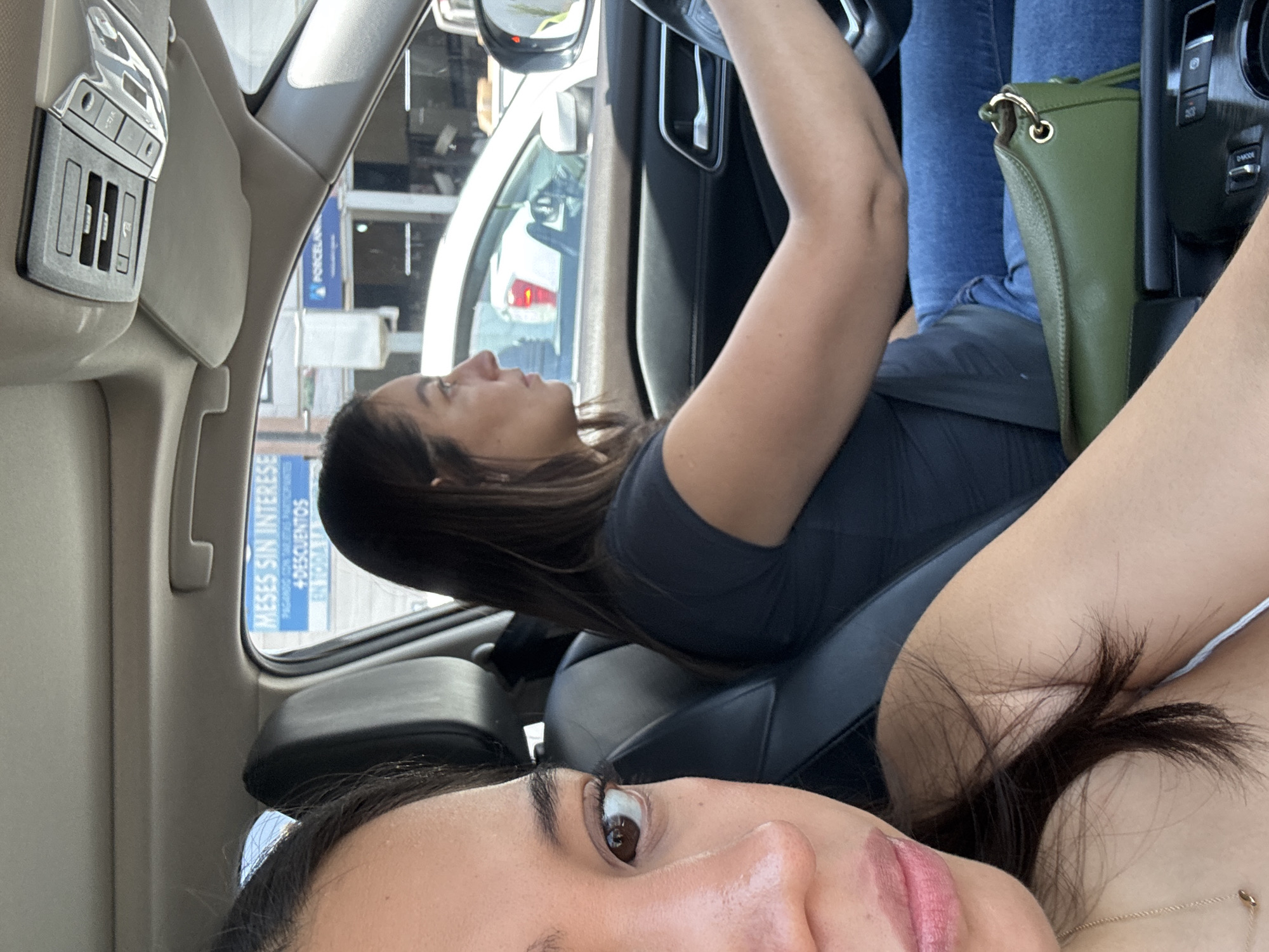 The image captures a casual moment inside a modern car during bright daylight. Two women are the main subjects. In the foreground, part of a woman's face is visible, including her right eye, nose, and mouth adorned with pink lipstick. She has dark hair and appears to be looking towards the camera, suggesting a selfie or a close-up shot.
Behind her, seated in the driver's seat, another woman is seen from the torso up. She has dark hair, wears a dark top and blue jeans, and is looking forward. Her arms are raised, likely engaged with the steering wheel, indicating she is driving or a passenger occupying the driver's seat.
A green handbag rests near the driver's lap. The car's interior features a light-colored ceiling and visible overhead console controls. Through the car windows, a sunny outdoor environment is apparent, with buildings and a white vehicle. A blue sign outside bears Spanish text: "MESES SIN INTERESES" (Months without interest), "+ DESCUENTOS" (+ Discounts), and partially "EN ORRA". This text confirms a Spanish-speaking location, aligning with the provided context of Puebla, Mexico. The overall atmosphere seems relaxed.
