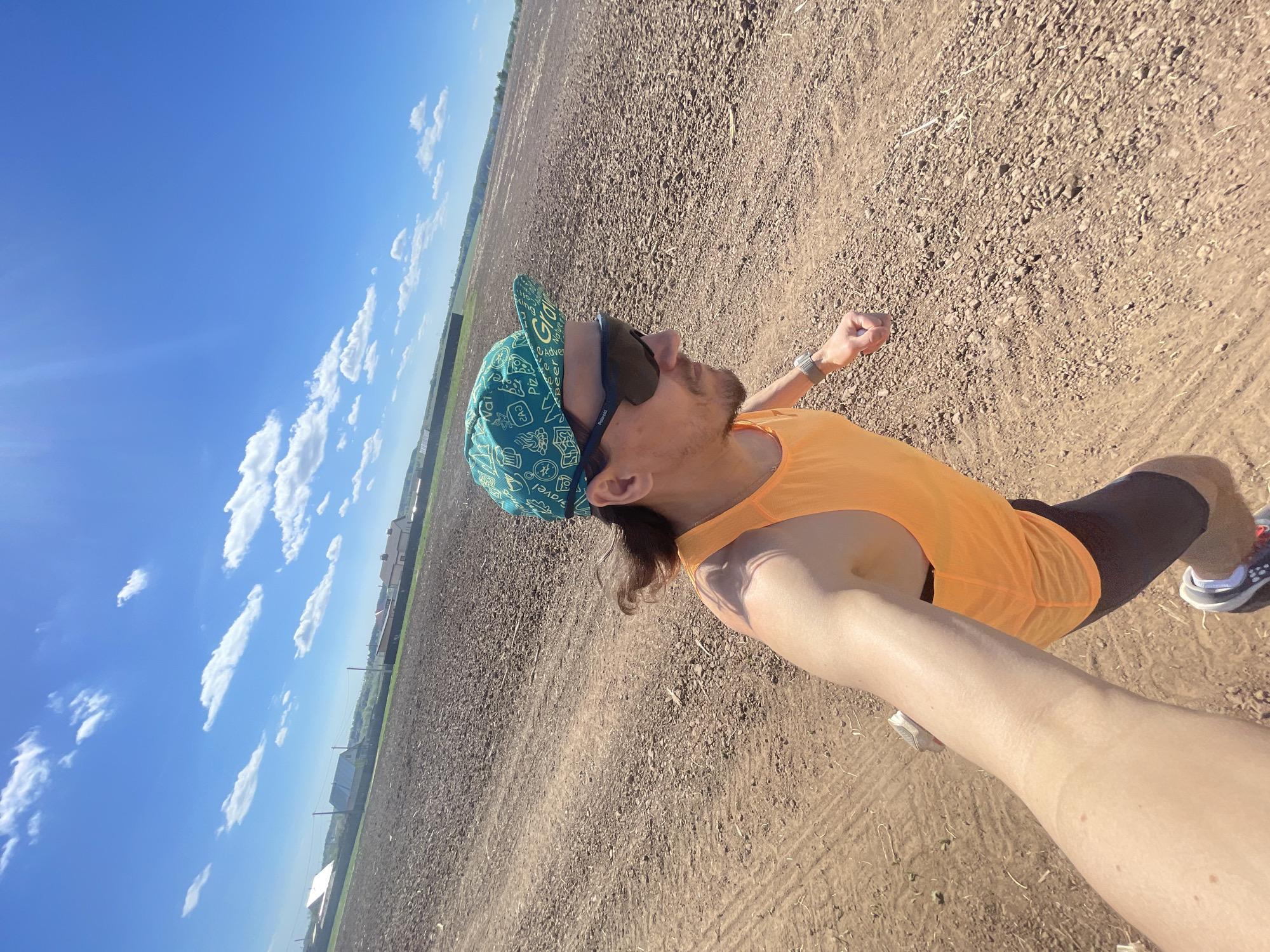 A ground-level outdoor shot captures a man in Перевозное, Russia, seemingly running. He is wearing a turquoise cap with assorted images, dark sunglasses, an orange sleeveless shirt, and dark athletic shorts. His hand holding the camera is visible, along with a watch on his wrist. The background is a vast, ploughed field under a blue sky dotted with scattered white clouds. A faint structure, possibly a building, is visible in the distance.