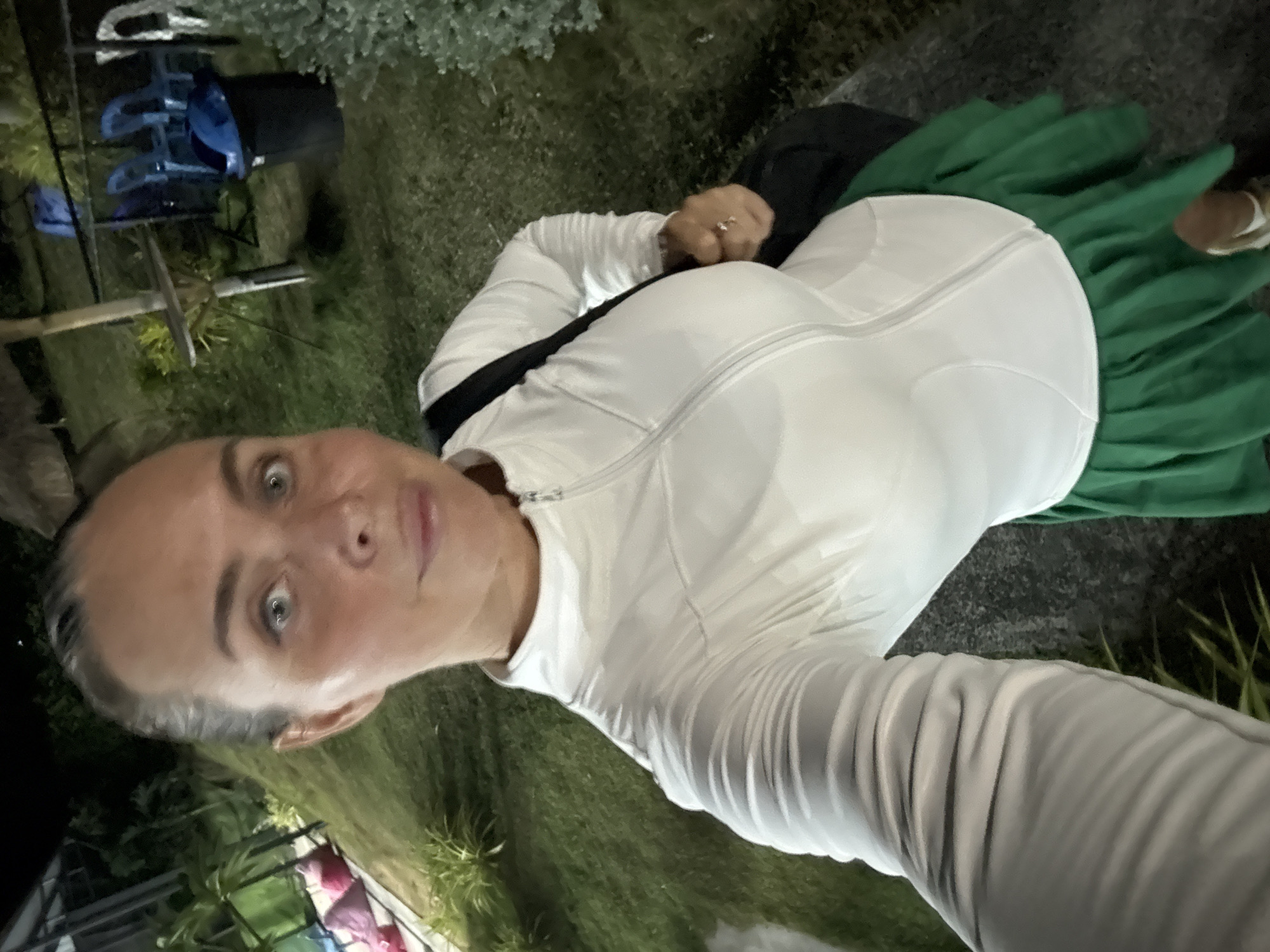 A woman is taking a selfie in a nighttime outdoor setting. She has light skin, light blue eyes, and her dark hair is pulled back. She is wearing a white zip-up jacket over a white sports bra and a green pleated skirt. She is also wearing a black bag over her right shoulder and a ring on her left hand. The background shows a grassy area with some plants, a table and chairs, and a trash can. The image appears to have been taken in the Ko Samui District of Thailand.