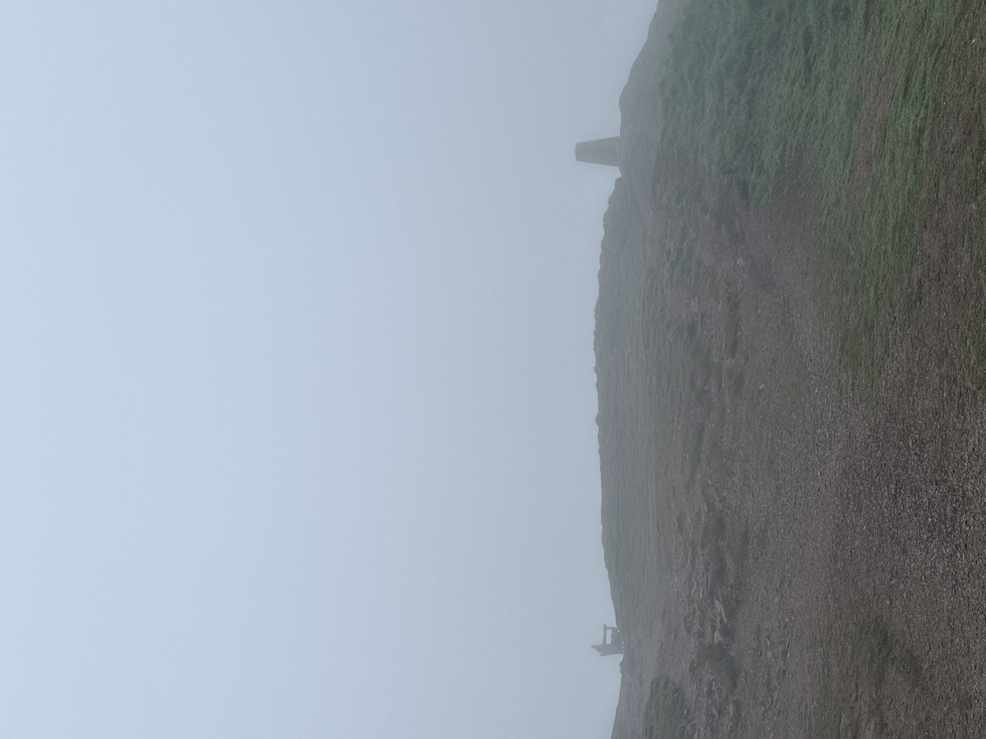 The image shows a mountain slope shrouded in heavy fog. The terrain is a mix of grassy patches and rocky, gravelly areas. A small structure, possibly a trig point or marker, is visible near the top of the slope. In the lower part of the image, a small platform or lookout point can be seen. The overall environment suggests a remote, mountainous location with limited visibility due to the fog.