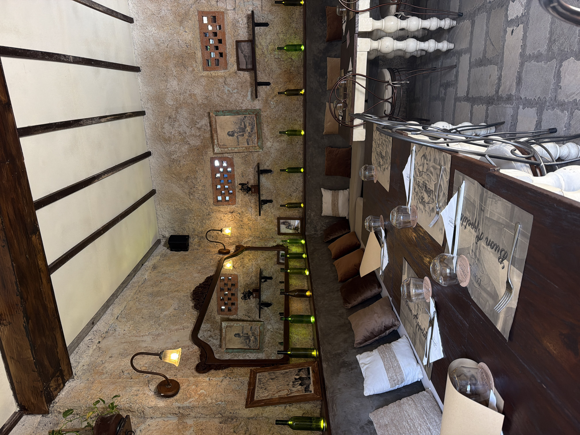 This is an indoor shot of a restaurant in Kuta Selatan, Indonesia. The interior is characterized by stone walls, decorated with vintage picture frames, wine bottles displayed on shelves, and small decorative lamps. The scene is focused on a long, dark wood table set for dining. Each place setting includes a placemat, glassware, silverware, and a folded napkin. Cushions serve as seating on one side of the table. The flooring appears to be made of stone. There are no people visible in the photograph.