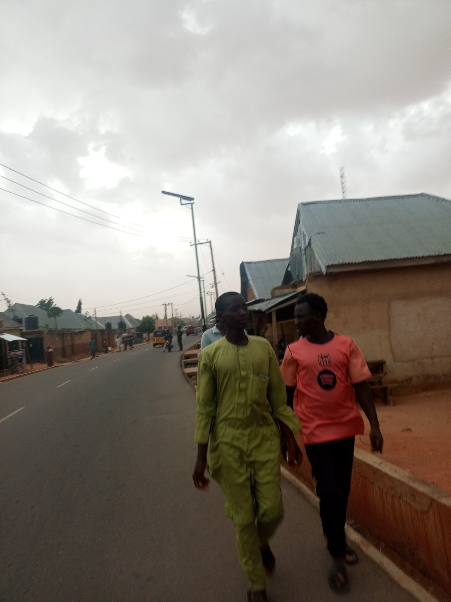 Two men are walking along a paved road in a city. The man on the left is wearing a light green long-sleeved shirt and matching pants, while the man on the right is wearing a salmon-colored short-sleeved shirt and black pants. Both are wearing sandals. They are walking on the edge of the road, next to a low concrete barrier.
In the background, there are houses with corrugated metal roofs. There are also utility poles and streetlights along the road. The sky is overcast.
The city appears to be of modest development, with basic infrastructure.