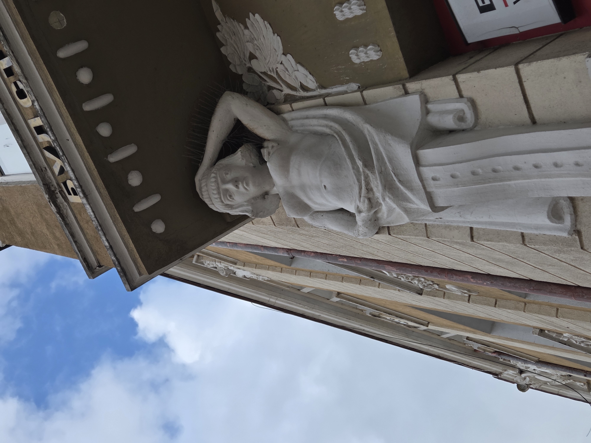 A low-angle shot depicts an architectural detail of a building in Varna, Bulgaria. A pale, sculpted female figure, likely a caryatid or atlas, supports an overhanging ledge. The figure is draped in fabric, and appears to be lying down. The background features a blue sky with scattered clouds. The architecture is characterized by beige-toned materials and decorative elements. A partial sign with red and white text is visible at the top right corner.