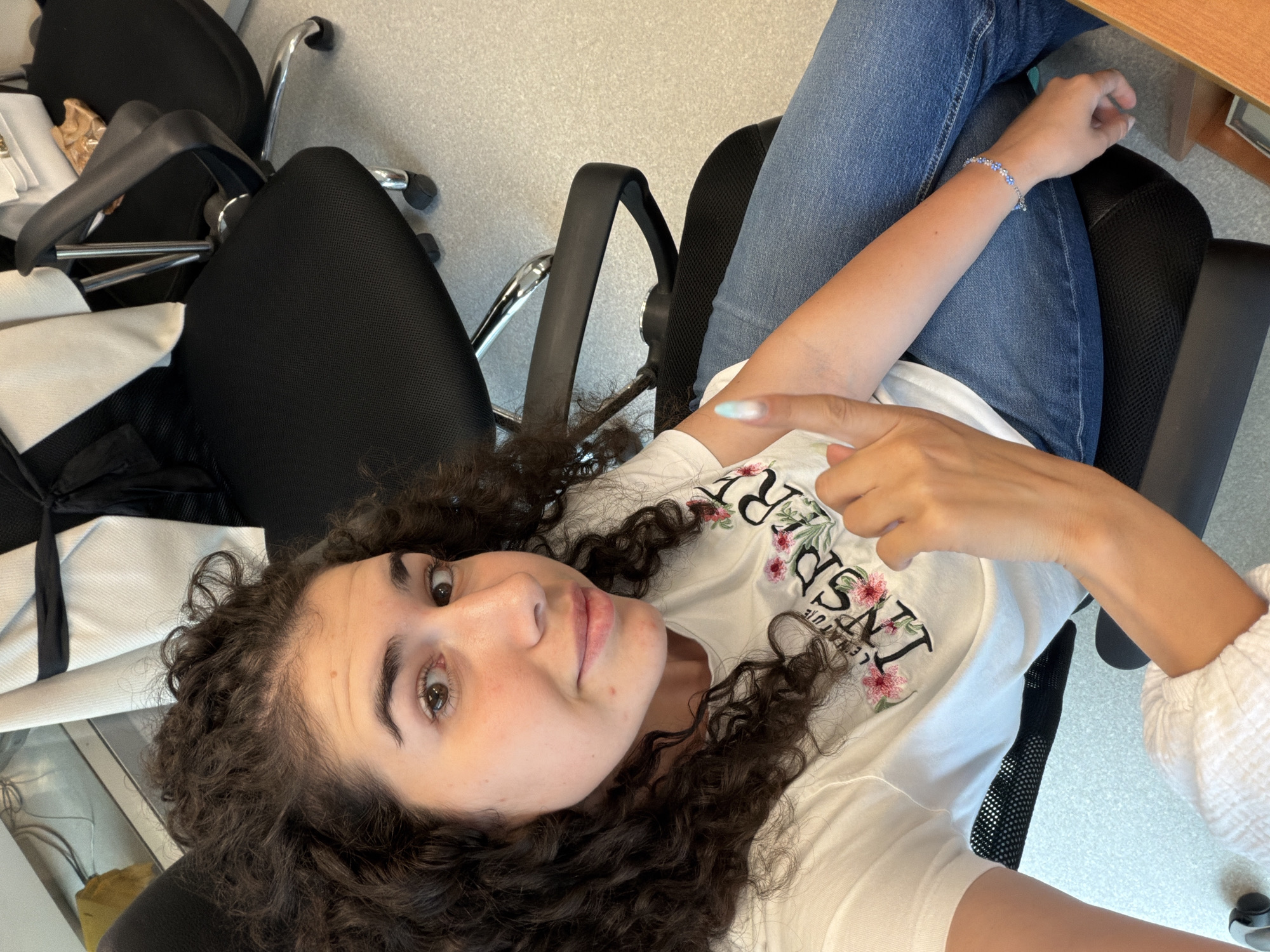 An indoor, high-angle, medium shot shows a young woman with long, dark curly hair sitting in a black office chair. She is wearing a white t-shirt with floral embroidery that reads "INSPIRE", and blue jeans. The woman is looking at the camera and holding up her right index finger, which has a light blue and white ombre nail polish. She is wearing a silver bracelet with blue beads on her left wrist. The background consists of other office chairs and a light gray speckled floor. The scene is an office environment.