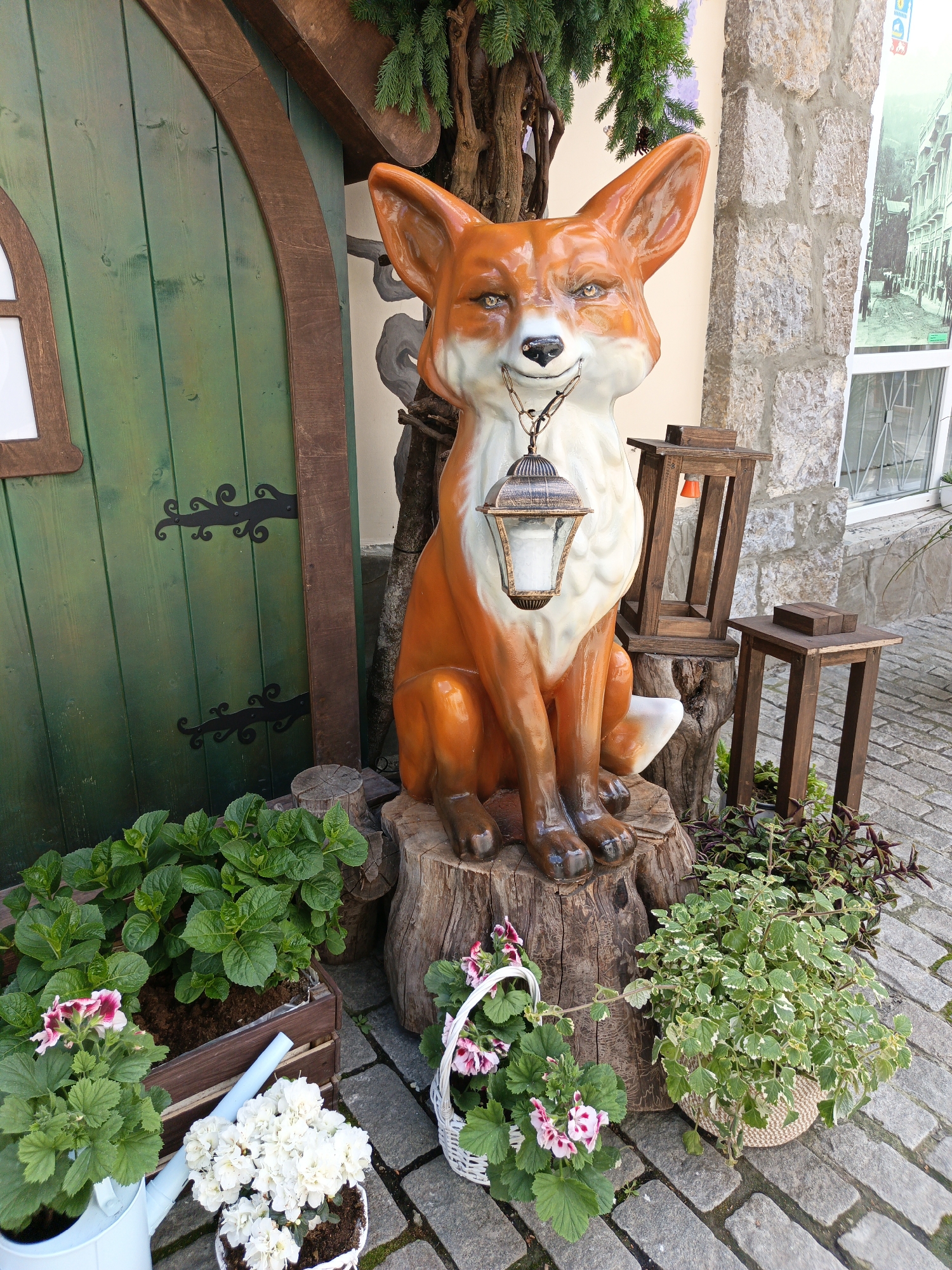 The image depicts a decorative scene outdoors. At the center, an orange fox statue with a white bib is perched on a tree stump, holding a lantern in its mouth with a chain. To the fox's left is a green hobbit-style door with decorative hinges, while to its right are two small wooden stools. A variety of potted plants surrounds the base, including white flowers, green leafy plants in a wooden box, and other leafy plants in wicker baskets. The scene appears to be located in Алупкинский городской совет, Ukraine, evident from the architectural style and the presence of cobblestone paving. The overall setting seems to be a decorated entrance or corner of a building.
