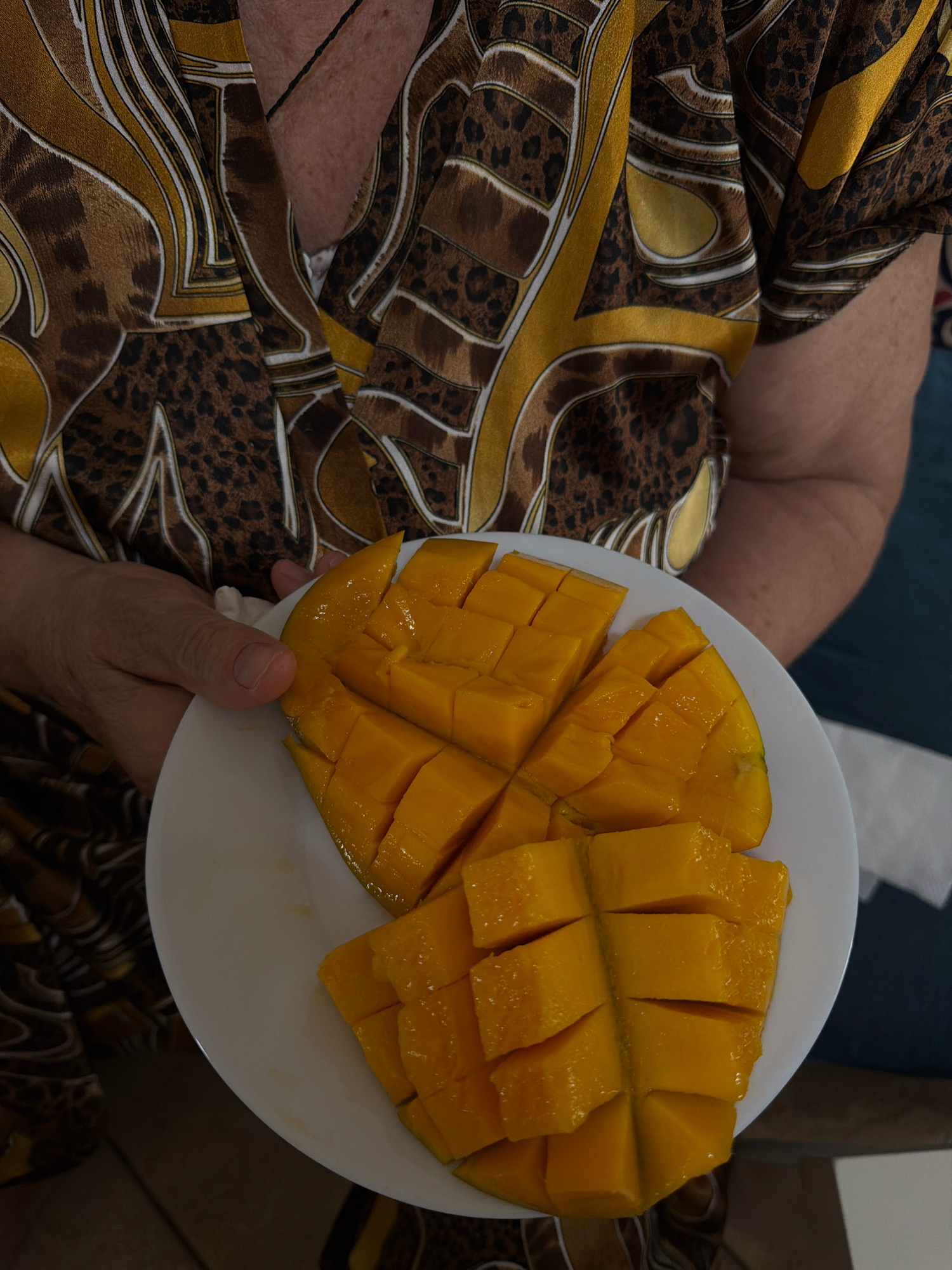 A person wearing a brown and gold patterned garment holds a white plate containing a sliced mango. The mango is cut into cubes, and its bright orange flesh is visible. The background shows a person's shoulder and neck, and a dark blue surface, possibly a chair or sofa. The image appears to have been taken indoors. There is no specific information in the image to determine the location within Sharm El Sheikh, Egypt.