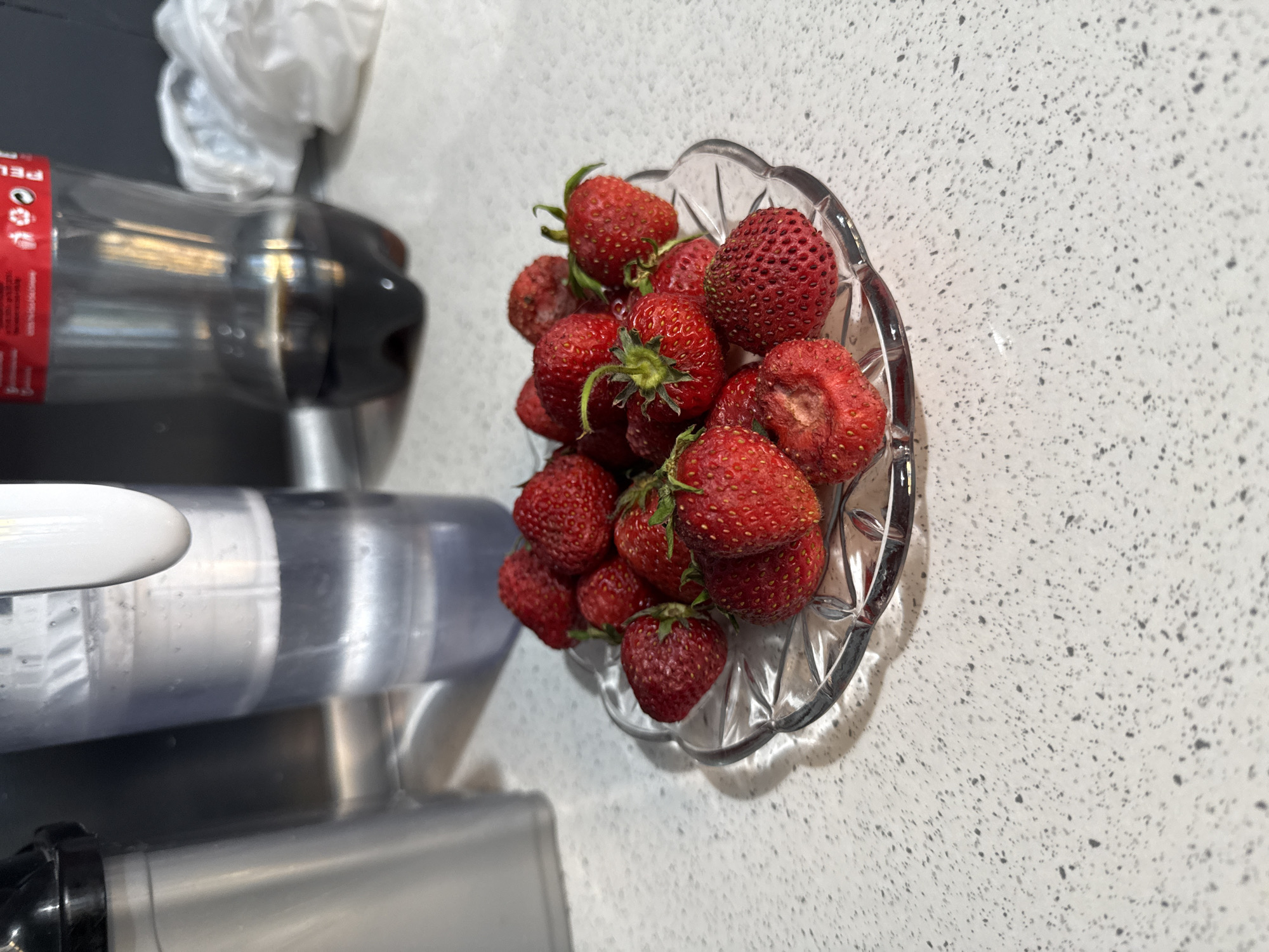 A clear glass decorative dish is filled with strawberries on a light speckled countertop. To the left is a beverage dispenser. In the background, there's a plastic bottle partially visible. The photo was taken indoors. There is no evidence to place the location within the city of Stara Zagora, Bulgaria.