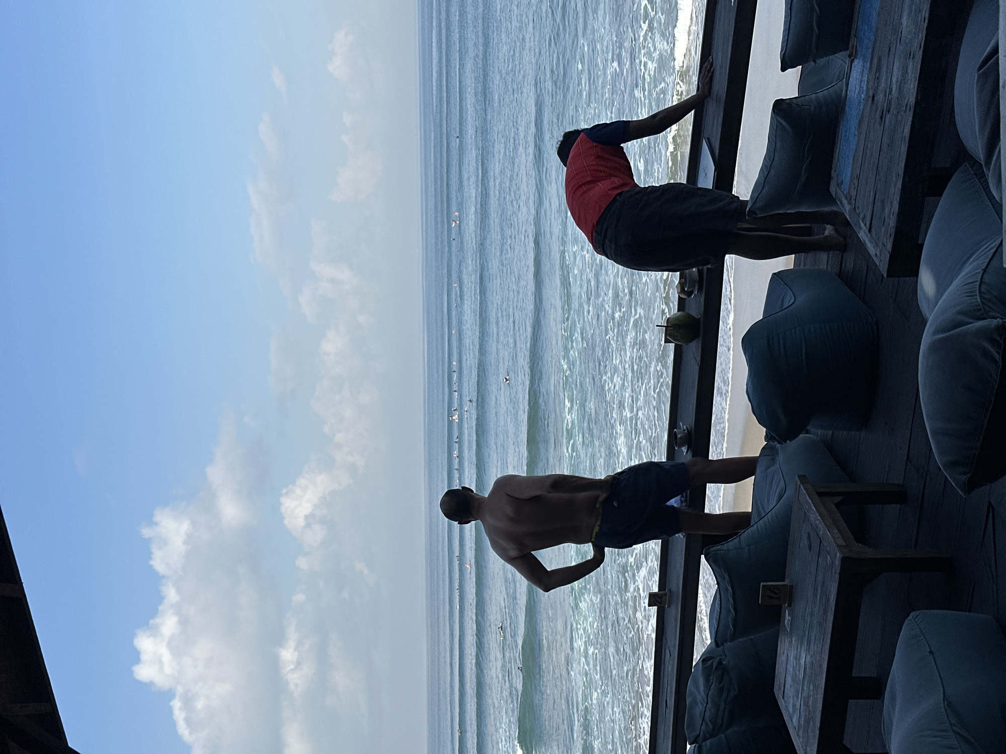 Two men are present at a beachside cafe in North Kuta, Indonesia, looking out at the ocean. The man on the left, shirtless and wearing dark blue shorts, stands with his hands on his hips, facing the sea. The man on the right, wearing a red shirt and dark shorts, is bent over, leaning on a wooden counter. Numerous surfers are visible in the water. In the foreground, there are several beanbag chairs and wooden tables, some of which have table numbers. A coconut and a cup of coffee are on the counter. The sky is blue with scattered clouds.