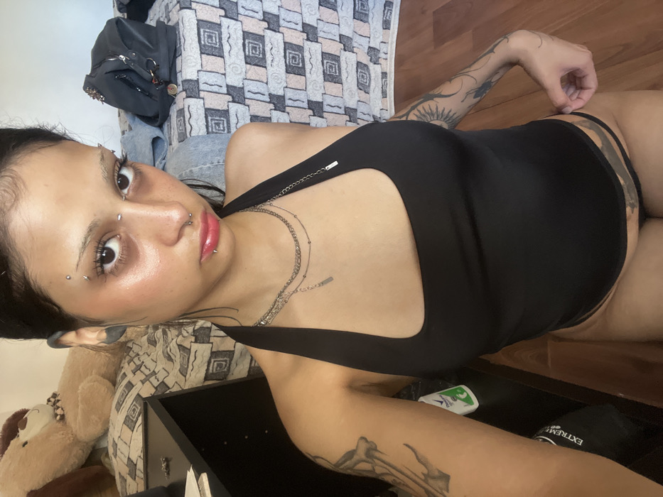 A woman lies on her side, facing the camera with a direct gaze. She is dressed in a black one-piece swimsuit or bodysuit. Visible piercings adorn her face, including the eyebrows, nose, and lips. Two silver necklaces accent her neckline. Tattoos are visible on both arms. The background features a bed with patterned bedding, a teddy bear, and a dark-colored piece of furniture with items like deodorant and a bottle. The lighting is soft, highlighting her skin.