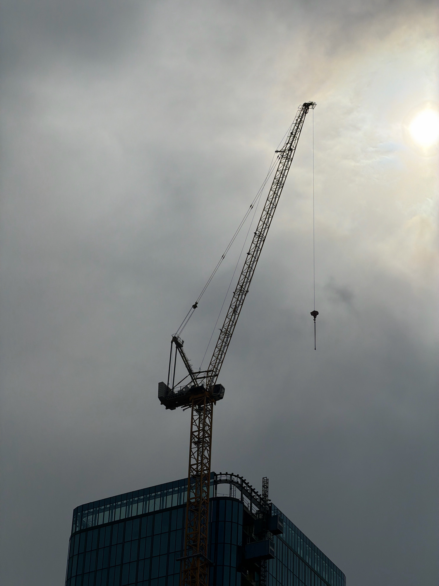 The image shows a construction crane silhouetted against a cloudy sky, with a bright sun partially visible in the upper right corner. The crane extends diagonally from the bottom left to the upper right of the frame, with its hook hanging down on a cable. Below the crane, the upper portion of a modern glass-faced building is visible. The scene appears to be a construction site in an urban environment. The location is identified as Wrocław, Poland. There are no people visible in the image. The overall mood is somewhat dramatic due to the lighting and the imposing presence of the construction equipment.