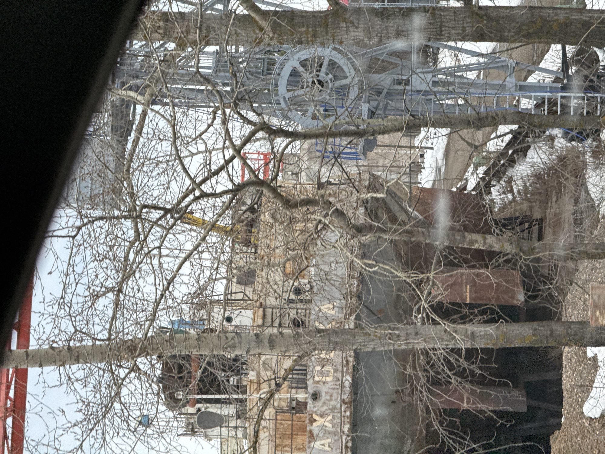 A view from inside a vehicle captures a snowy scene in Chaikovsky, Russia, where industrial structures are partially obscured by bare tree branches. Prominent among the structures is a large gray metal wheel mechanism mounted on a framework. A multi-story building with a sign, likely industrial or commercial, is visible further down. Snow covers parts of the ground and nearby structures, contrasting with the darker colors of the building and tree branches. The presence of the tree branches suggests an urban area where nature intertwines with industrial elements.