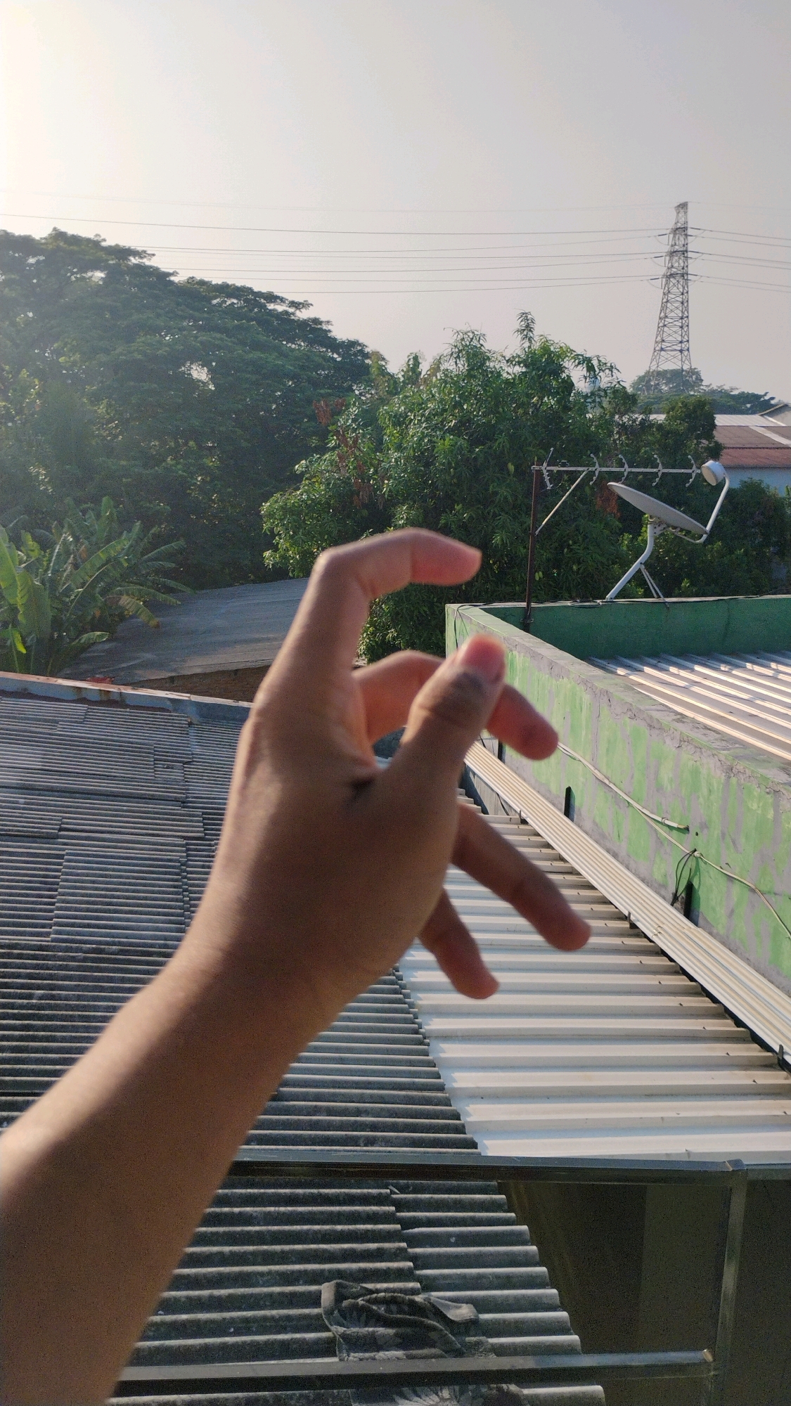A person's hand is in the foreground, obscuring a portion of the image. The hand is positioned in front of two sloped rooftops. The rooftop closest to the viewer is covered with corrugated metal. Beyond this, the second rooftop is also metal, but appears to be painted white or a very light color. A green structure runs along the edge of the second rooftop. In the background, there are several trees and a tall electrical tower. The sky is a pale, hazy blue, suggesting it may be early morning or late afternoon. The scene appears to be an outdoor urban or suburban environment. The location in Gumpang, Indonesia, is not discernible from the visual information alone.
