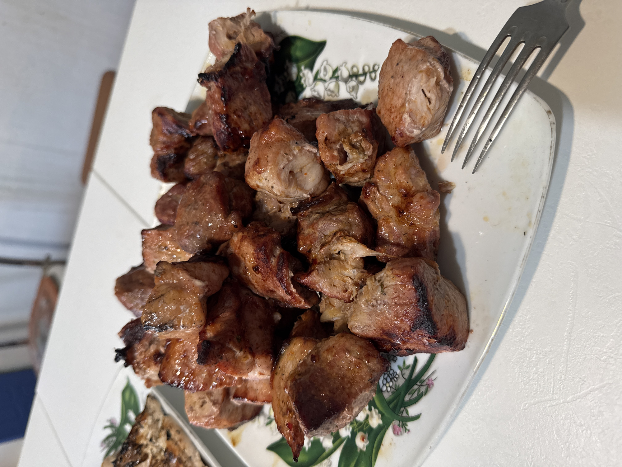 A close-up, high-angle shot shows a white rectangular plate filled with grilled meat cubes. The meat is various shades of brown, with some pieces exhibiting char marks. The plate has a decorative pattern of green leaves and small white flowers along one edge. To the right of the plate, a silver fork rests on the white surface of a table. In the background, the blurry image of a chair and part of a wall can be seen. The setting appears to be a domestic dining area. The location in the city of Бигила, Russia, is unknown.