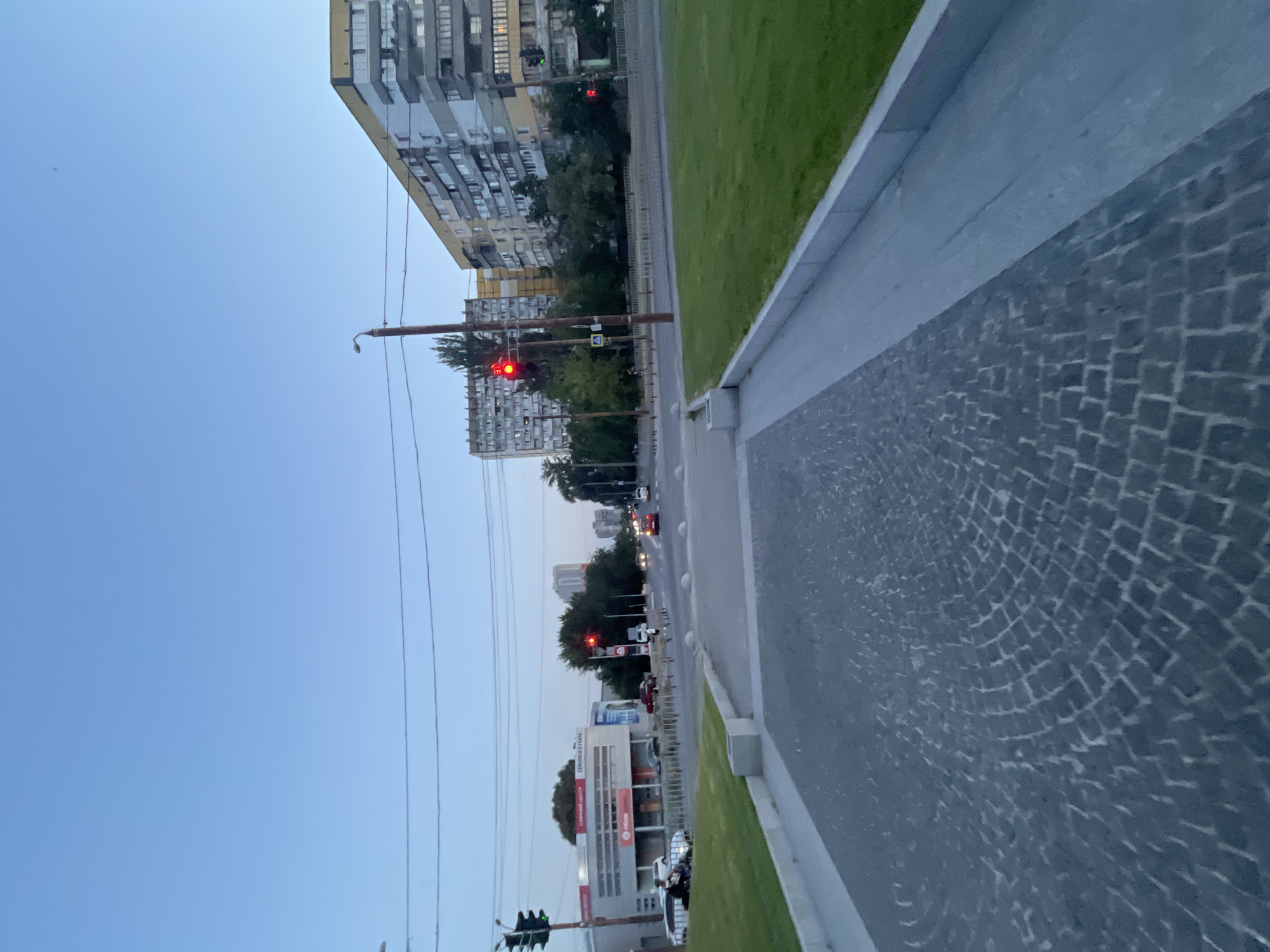 The image depicts a city street in Dnipro, Ukraine, at dusk. The perspective is from a low angle, looking forward down a cobblestone path towards an intersection with a multi-lane road. Several cars are visible on the road, some stationary at a red traffic light, while others are moving. Two prominent apartment buildings with multiple floors and balconies are on the right side of the street, one yellow and gray, the other predominantly gray. Across the intersection, a long, single-story building with the Bridgestone logo is visible. Traffic lights are red in several locations. There are trees and utility poles lining the street. A grassy area with a concrete curb runs alongside the cobblestone path in the foreground. The sky is a clear, pale blue, indicative of twilight. No individuals are clearly identifiable or engaged in significant actions.