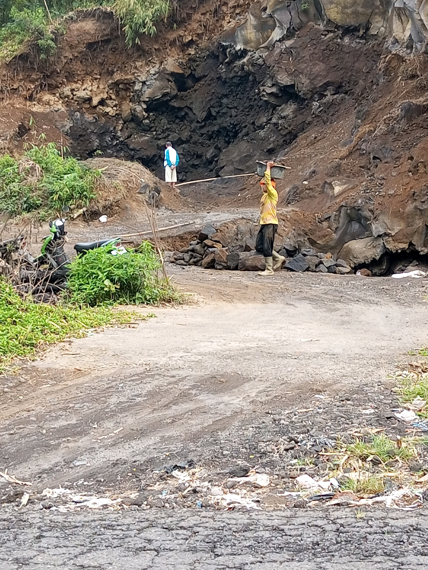 In Andongsili, Indonesia, a dirt road winds through a rocky hillside with sparse vegetation. Two individuals are visible. One person, wearing a blue and white top and light-colored trousers, stands near the base of the hill. Further down the road, another person is carrying a flat object, possibly a tray or basket, balanced on their head, dressed in a yellow long-sleeved shirt and dark pants, with light-colored boots. A motorcycle is parked on the left side of the road, partially obscured by green foliage. The ground is uneven, with scattered rocks and debris, including some white plastic. The environment appears to be a work site or an area undergoing some form of construction or excavation.