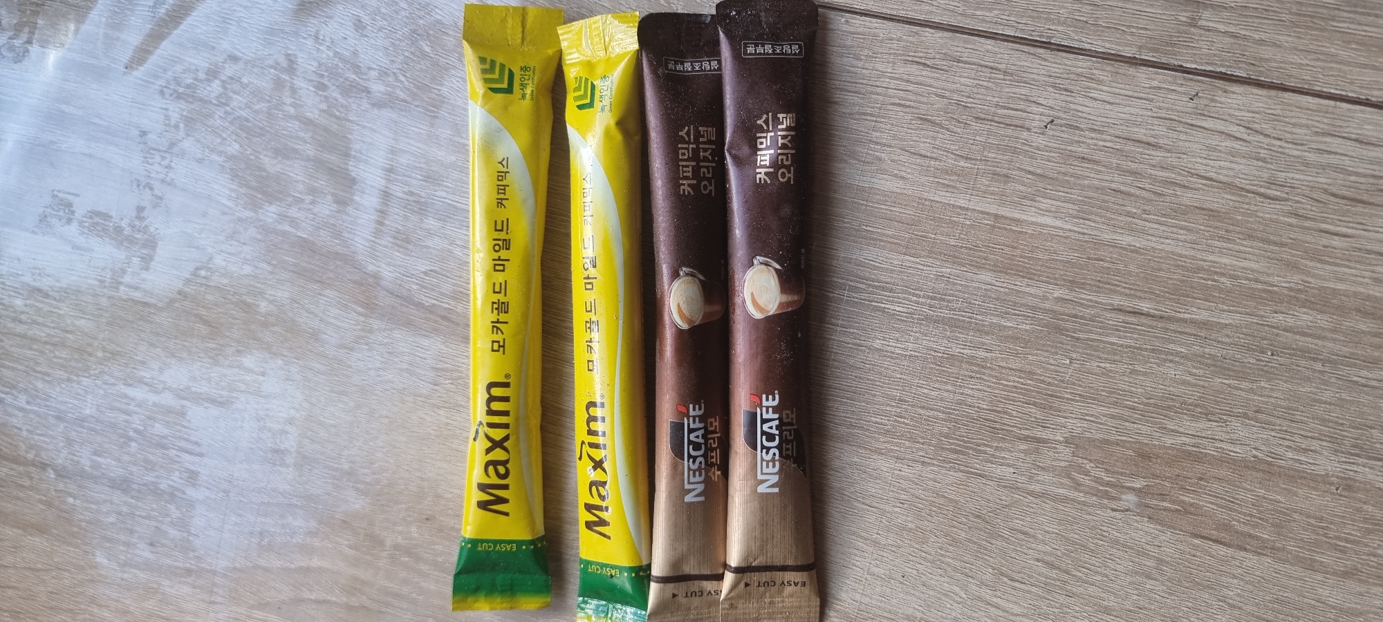 The image shows four individual coffee stick packets arranged horizontally on a light-colored wooden surface. From top to bottom, the packets are:
1. A yellow packet with "Maxim" logo and Korean text "모카골드 마일드 커피믹스". It also has a green certification logo on the right.
2. Another yellow packet, identical to the first, with the "Maxim" logo and Korean text "모카골드 마일드 커피믹스". It also has a green certification logo on the right.
3. A dark brown packet with the "NESCAFÉ" logo and Korean text "수프리모". An illustration of a coffee cup with foam is present on the right side.
4. A dark brown packet, identical to the third, with the "NESCAFÉ" logo and Korean text "수프리모". An illustration of a coffee cup with foam is present on the right side.
The wooden surface appears to be a floor with visible wood grain. The lighting is natural, casting a slight shadow from the packets onto the surface.
There is no information or visual evidence in the image to suggest it is located in the city of Aktobe, Kazakhstan. The description is purely based on the visual content of the image.