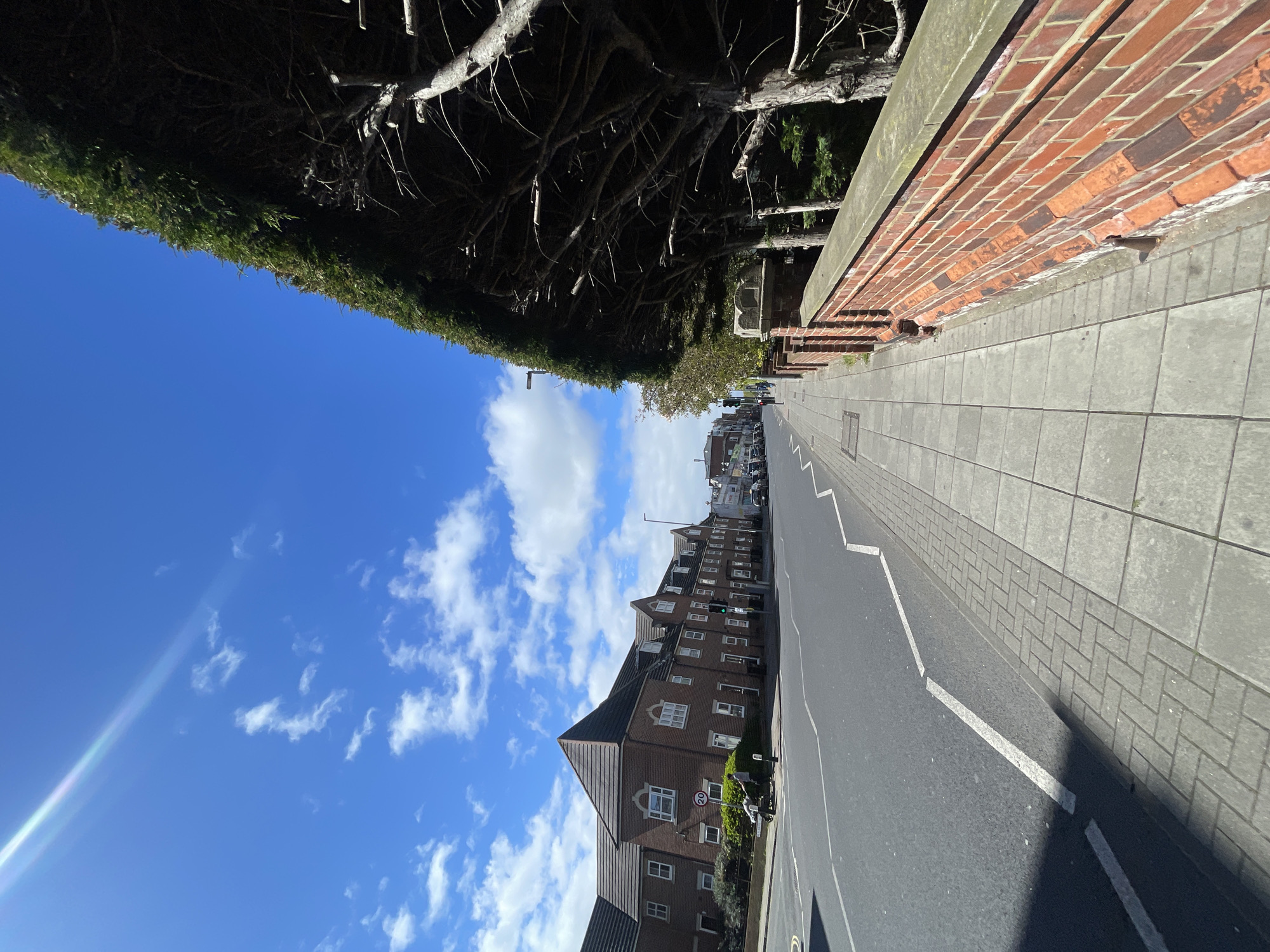 The image shows a street scene in Portsmouth, United Kingdom, under a partly cloudy sky. The perspective is from a low angle, looking down the length of the road.
On the right side of the road is a red brick wall, with a paved grey sidewalk adjacent to it. The road itself is asphalt and has white road markings, including what appears to be a broken line marking. A terraced housing structure with brickwork architecture, grey roof, and white windows is on the left side.
There is a street sign visible on the left side of the road. Vehicles are parked further down the street.
Overhanging the left side of the road are dense bushes and the branches of a tree. The sky is blue with scattered white clouds. A lens flare is visible in the sky on the left. The overall lighting is bright, suggesting a sunny day.