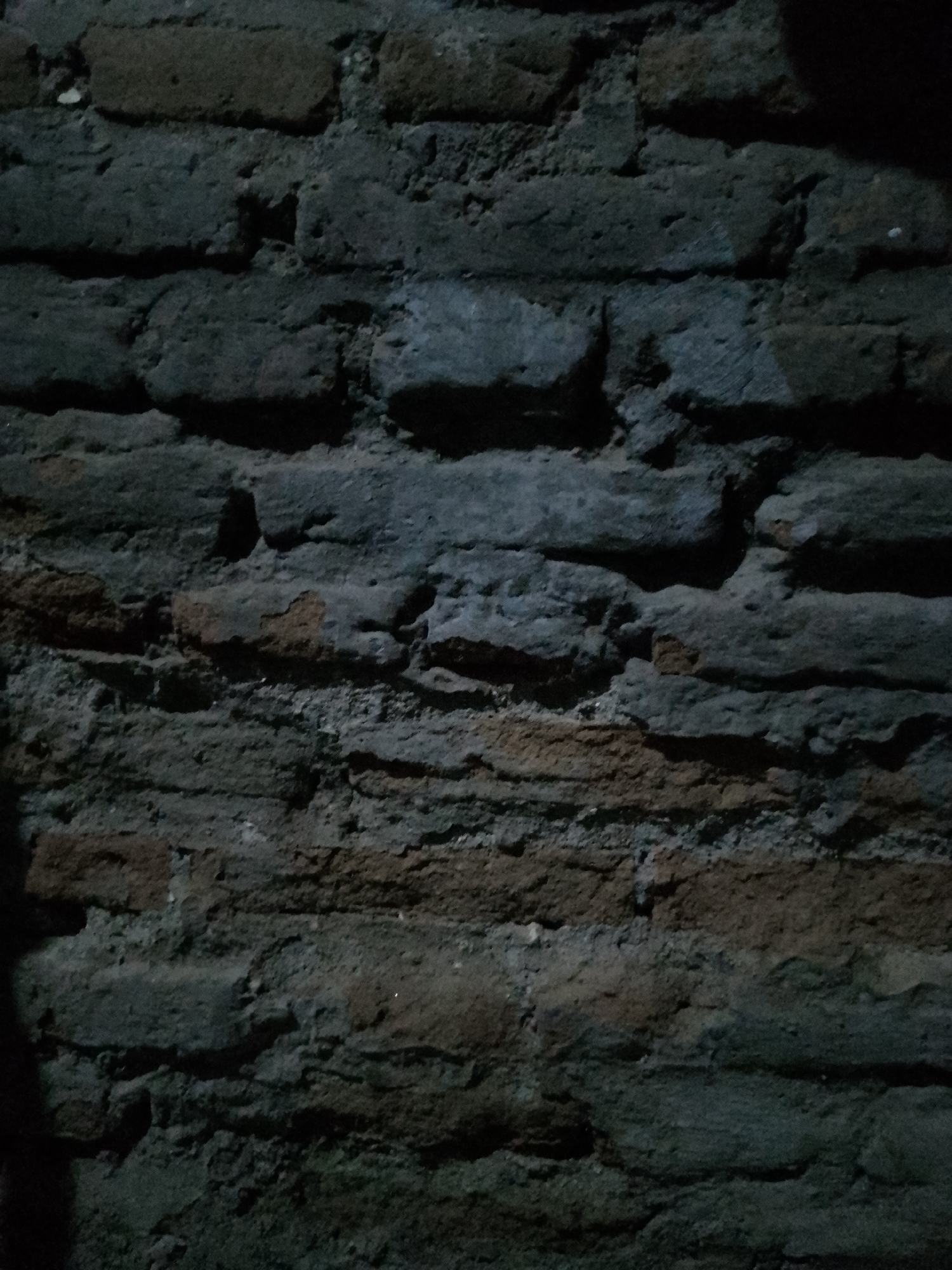 The image shows a close-up, dark, and textured view of an old brick wall. The bricks are rough, uneven, and appear to be aged, with some areas showing mortar that is chipped or missing. The color palette is dominated by dark grays, blues, and some earthy brown tones from the bricks. The lighting is dim, creating strong shadows that emphasize the texture and imperfections of the wall. The overall impression is one of age and weathering. The location in Kauman, Indonesia, is suggested, but the image itself does not contain any explicit visual cues to confirm this specific location; it only depicts a generic brick wall. There are no people, animals, or specific objects visible, only the distressed surface of the wall.