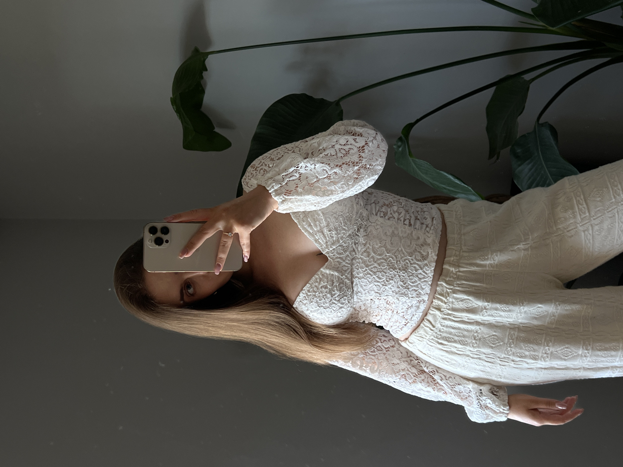 A person is taking a selfie in a mirror. The person has long blonde hair and is wearing a white lace top with long sleeves and white patterned pants. They are holding a smartphone in their right hand. In the background, there is a large green plant with broad leaves. The setting appears to be indoors, with a gray wall and a reflective surface, likely a mirror, that shows the person and the plant. The lighting suggests it could be daytime. The image is oriented sideways.