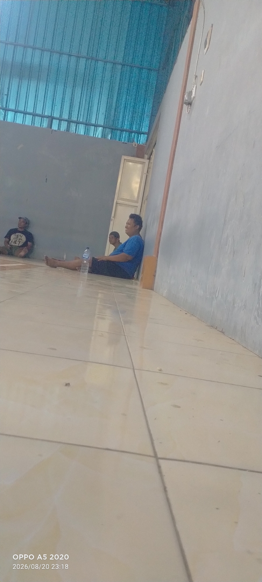 Three individuals are seated on a tiled floor indoors. The person on the left is wearing a black t-shirt and a hat and is looking upwards. The person in the middle is wearing a blue t-shirt and black pants, and is looking towards the right. The person in the background is wearing a blue t-shirt and also appears to be looking towards the right. A water bottle is placed on the floor next to the middle person. The environment appears to be a covered outdoor area with a corrugated blue roof and a gray wall. The image was taken on August 20, 2026, at 23:18.