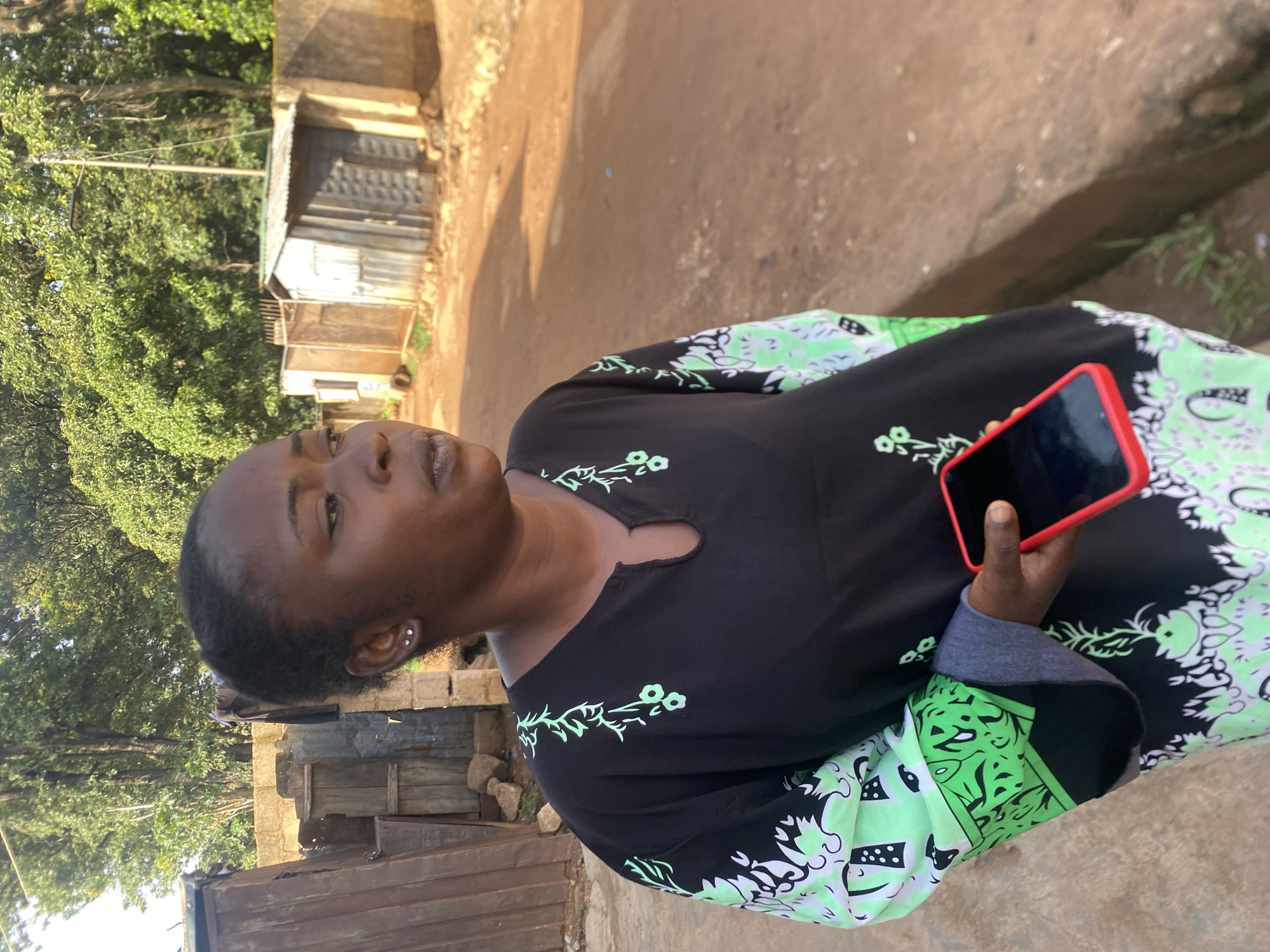 A medium shot captures a Black woman standing outdoors on a dirt road in Bukuru, Nigeria. She is facing to her right, looking away from the camera with a neutral expression. She has her hair pulled back and wears small silver stud earrings. Her attire consists of a black, loose-fitting, long-sleeved garment adorned with a green and white floral pattern on the sleeves and hem. She is holding a smartphone with a bright red case in her right hand, which is positioned in front of her body.
The background suggests a semi-urban or rural setting. To the left, there are structures made of corrugated metal and wooden planks, appearing somewhat makeshift. A brick wall is visible behind these structures. The road itself is unpaved and reddish-brown, with some scattered debris. Beyond the road, more corrugated metal structures are visible, resembling small shops or dwellings. Lush green trees and foliage line the background, indicating a natural environment bordering the built-up area. The lighting suggests it is daytime, with sunlight casting shadows.