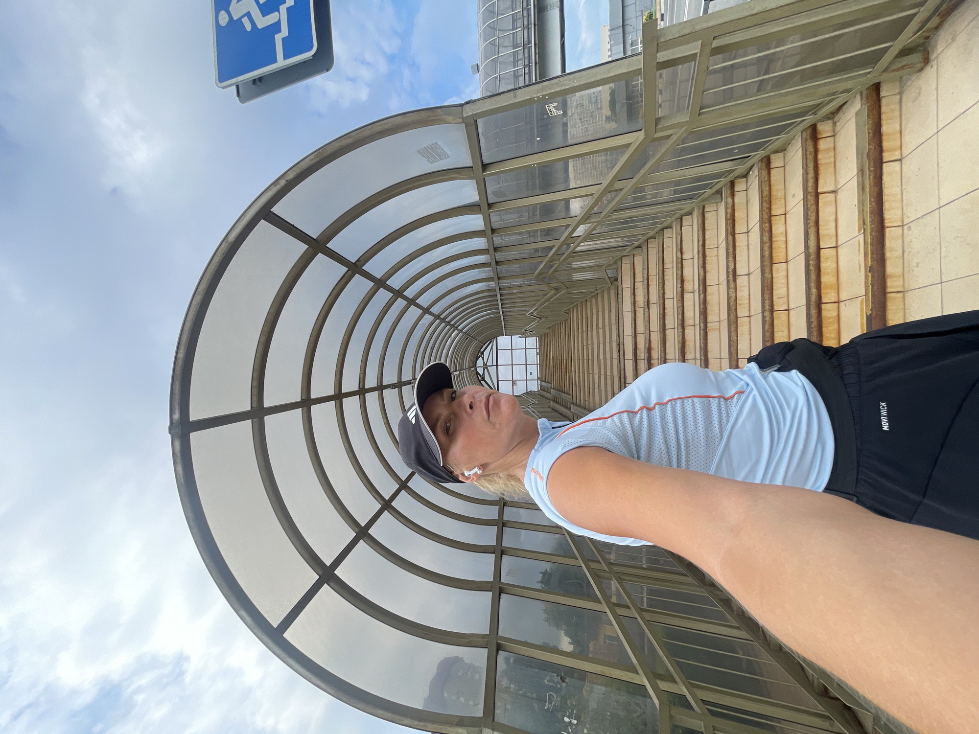 A woman is taking a selfie outdoors in a covered walkway. She is wearing a light blue athletic shirt with orange trim, black shorts, a black baseball cap, and white wireless earbuds. Her expression is neutral, and she appears to be looking directly at the camera.
The walkway has a curved, translucent roof supported by metal beams. To the right, there are steps with light-colored tiles and rusted metal railings. In the background, to the left and upper background, there are buildings and the sky, which is partly cloudy. A blue sign with a white symbol indicating accessibility for people with disabilities is visible in the upper left corner of the frame. The lighting suggests it is daytime, and the weather appears to be mild. The image is oriented sideways. Text visible on the shorts reads "MOVİWİCK".