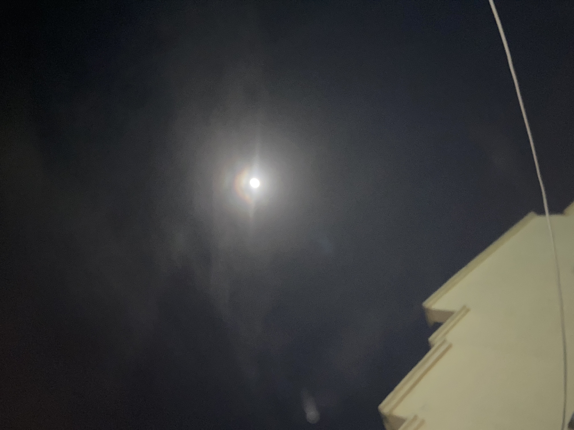 The image shows a night scene in Lagos, Nigeria. The sky is dark with some clouds, and a bright full moon is visible with a faint halo around it. In the lower right corner of the image, part of a light-colored building is visible, with a vertical cable or wire running alongside it. No people are visible in the image.