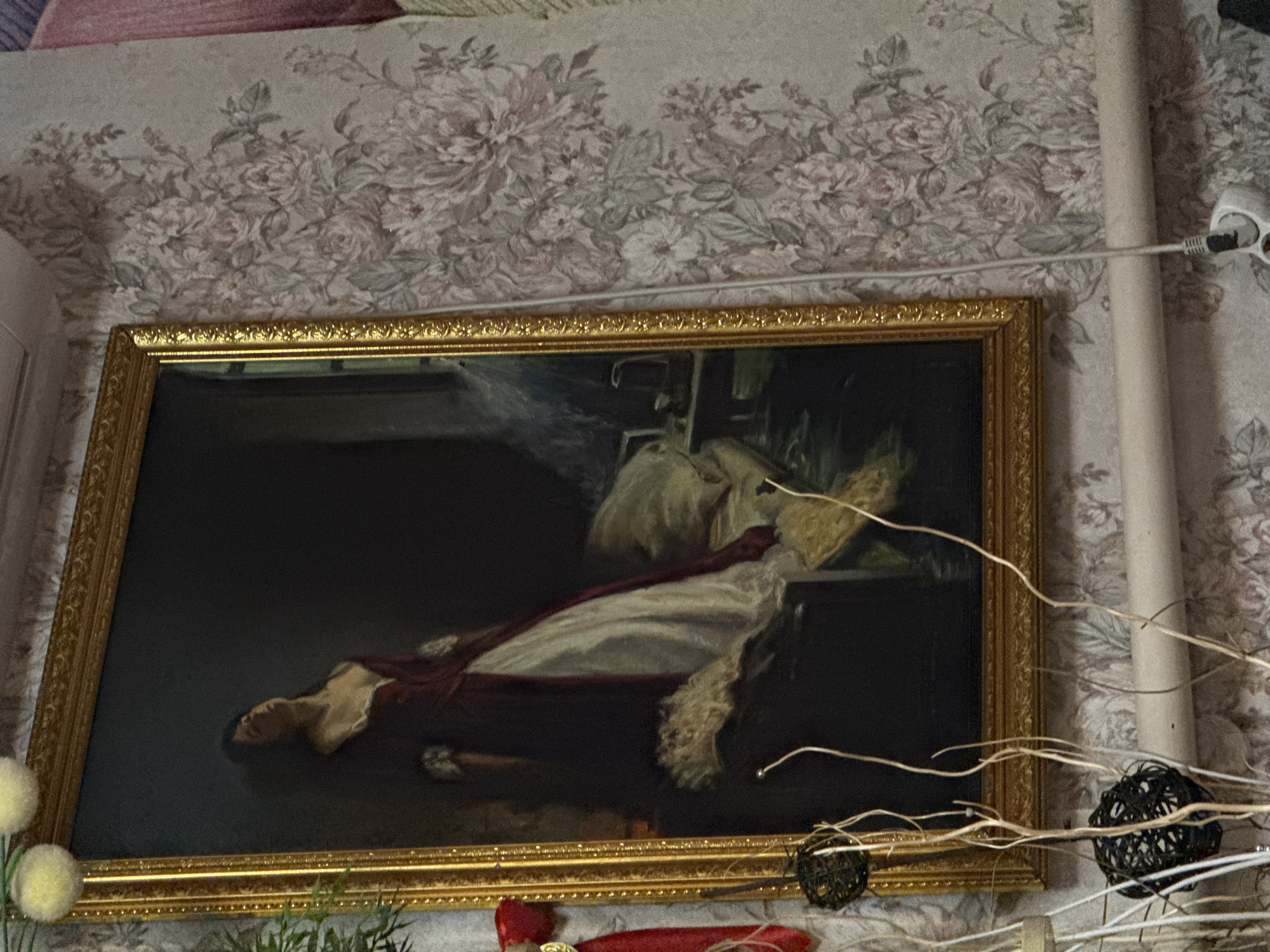 The image shows a framed painting hanging on a floral patterned wall in a seemingly indoor environment. The painting depicts a woman reclined and draped in fabric. Other visible objects include a power outlet cord, decorative branches, a fuzzy ball, and small red objects below the painting. No city is visible and the location is unknown.