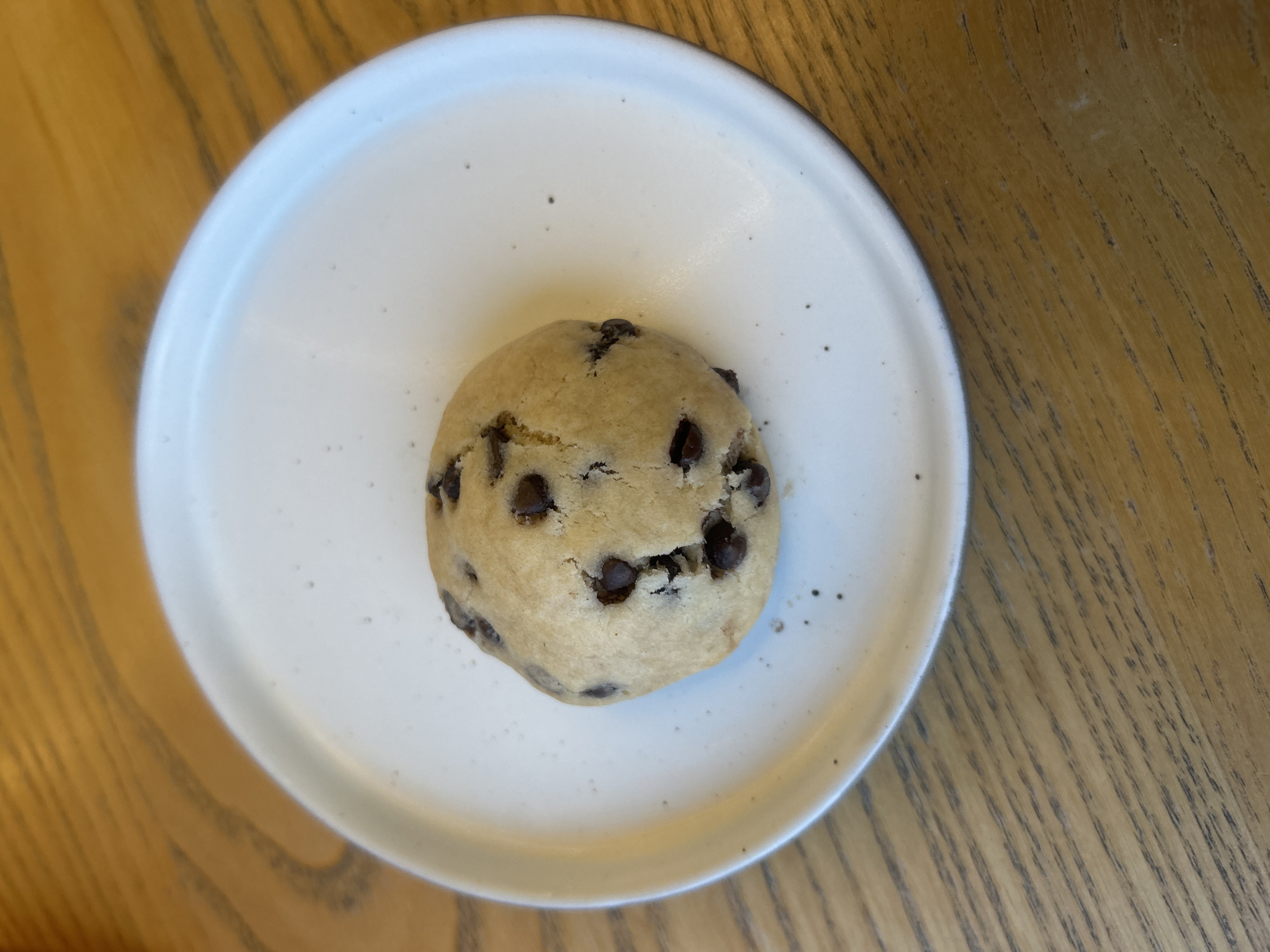 A chocolate chip cookie rests on a white plate, which sits on a wooden table. The scene is likely indoors.