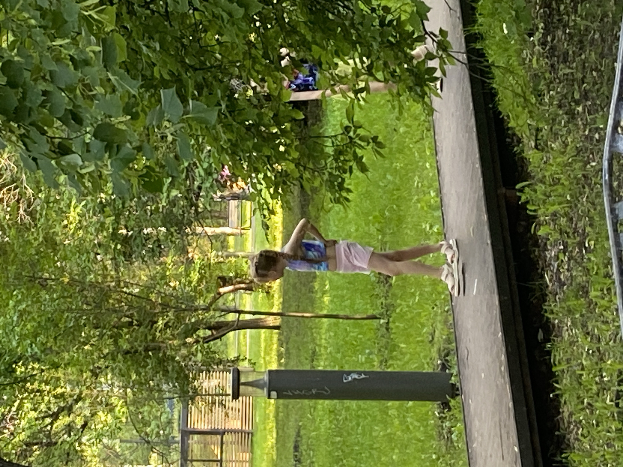 The image depicts a young girl with blonde braided hair standing on a paved walkway in a park-like setting. She is wearing a colorful tank top and light pink shorts, with white sandals on her feet. Her hands are on her hips, and she is looking to her left. To her left is a dark grey cylindrical object with a light on top, likely a lamp post. The background is filled with lush green grass and trees, some of which are casting shadows. In the distance, there are some buildings or structures with horizontal elements and more trees. The lighting suggests it is daytime, possibly late afternoon or early morning due to the soft light. The environment appears to be a public park or recreational area in Yekaterinburg, Russia, given the provided context.