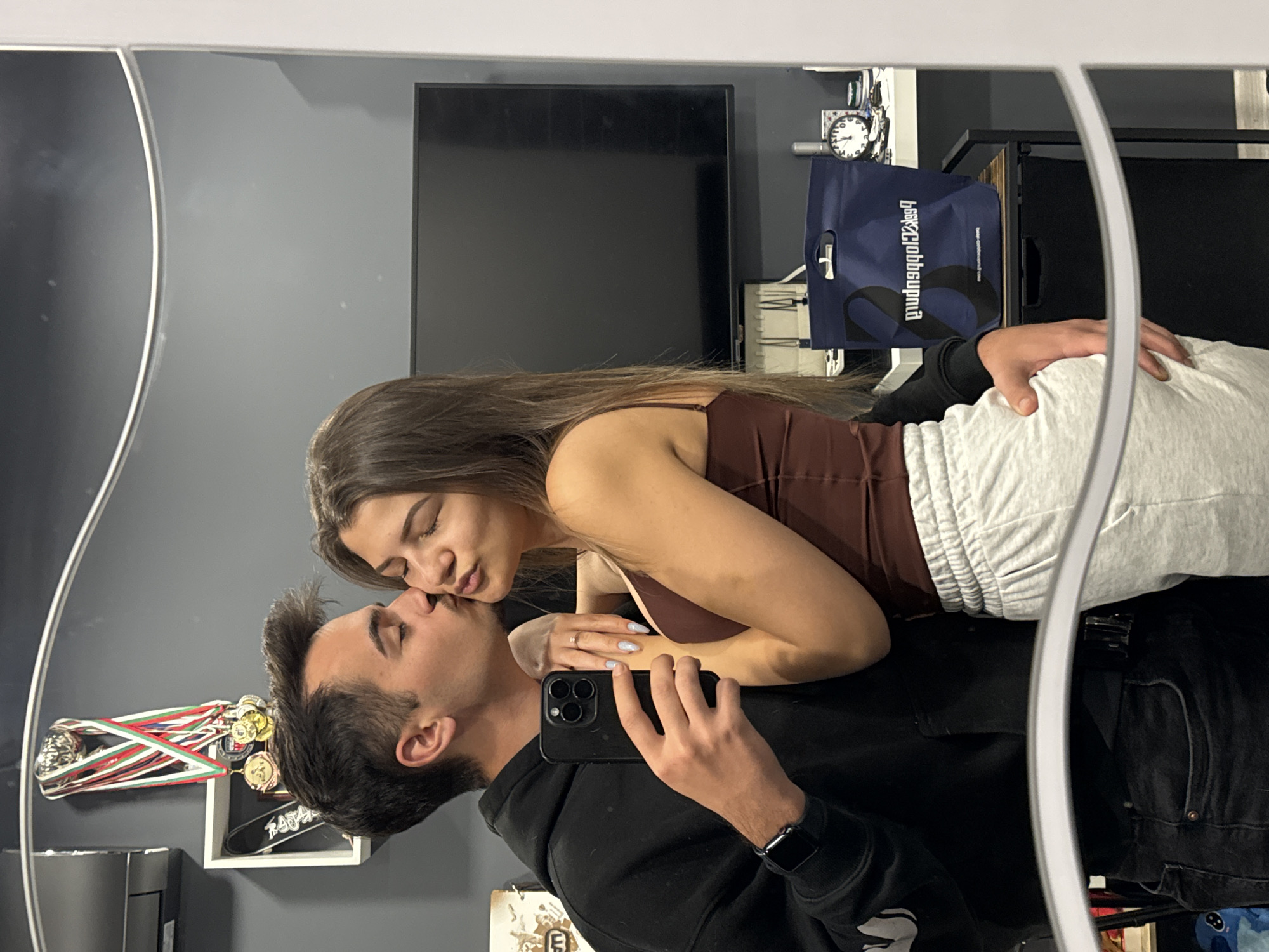 A young couple is seen in a mirror selfie indoors. The woman has long brown hair and wears a brown spaghetti-strap top and light gray sweatpants. The man wears a black hooded sweatshirt and dark jeans. He holds a black smartphone with three camera lenses and a silver wristwatch on his left wrist. They are kissing, with the woman's face tilted towards the man's. Behind them is a gray wall with a flat-screen television mounted on it. To the right of the TV is a blue shopping bag, a clock, and some other items on a shelf. To the left of the TV are some medals and a small shelf. The location is likely a private residence in Sofia, Bulgaria, based on the possible Bulgarian flag colors among the medals and general interior design.