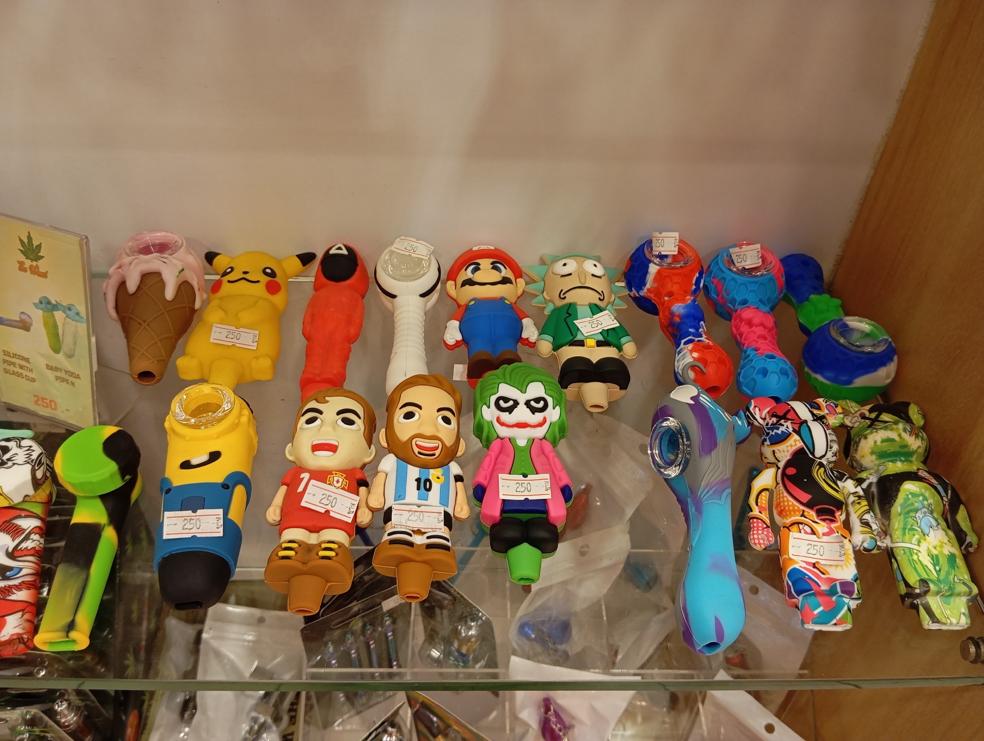 A shelf in a shop displays a variety of silicone pipes and accessories, primarily characterized by their novelty designs. The items are arranged in rows on a glass shelf against a light-colored wall and a wooden partition on the right.
In the top row, from left to right, are:
- A pink ice cream cone-shaped pipe.
- A yellow Pikachu pipe.
- An orange Squid Game mask figure.
- A white mushroom-shaped pipe.
- A figure resembling Super Mario.
- A figure resembling Rick Sanchez from Rick and Morty.
- Several colorful, spherical silicone attachments, some with pipe stems.
The bottom row features:
- A pipe with a black and green tie-dye design.
- A yellow Minion-shaped pipe.
- A figure of a soccer player with the number 7.
- A figure of a soccer player with the number 10.
- A figure of the Joker from Batman.
- A blue and purple pipe with a swirling marbled design.
- Two stylized bear-shaped figures with complex, colorful patterns.
Several items have small white tags with prices, reading "250". The overall lighting suggests an indoor retail environment. The shop is located in Karon, Thailand.