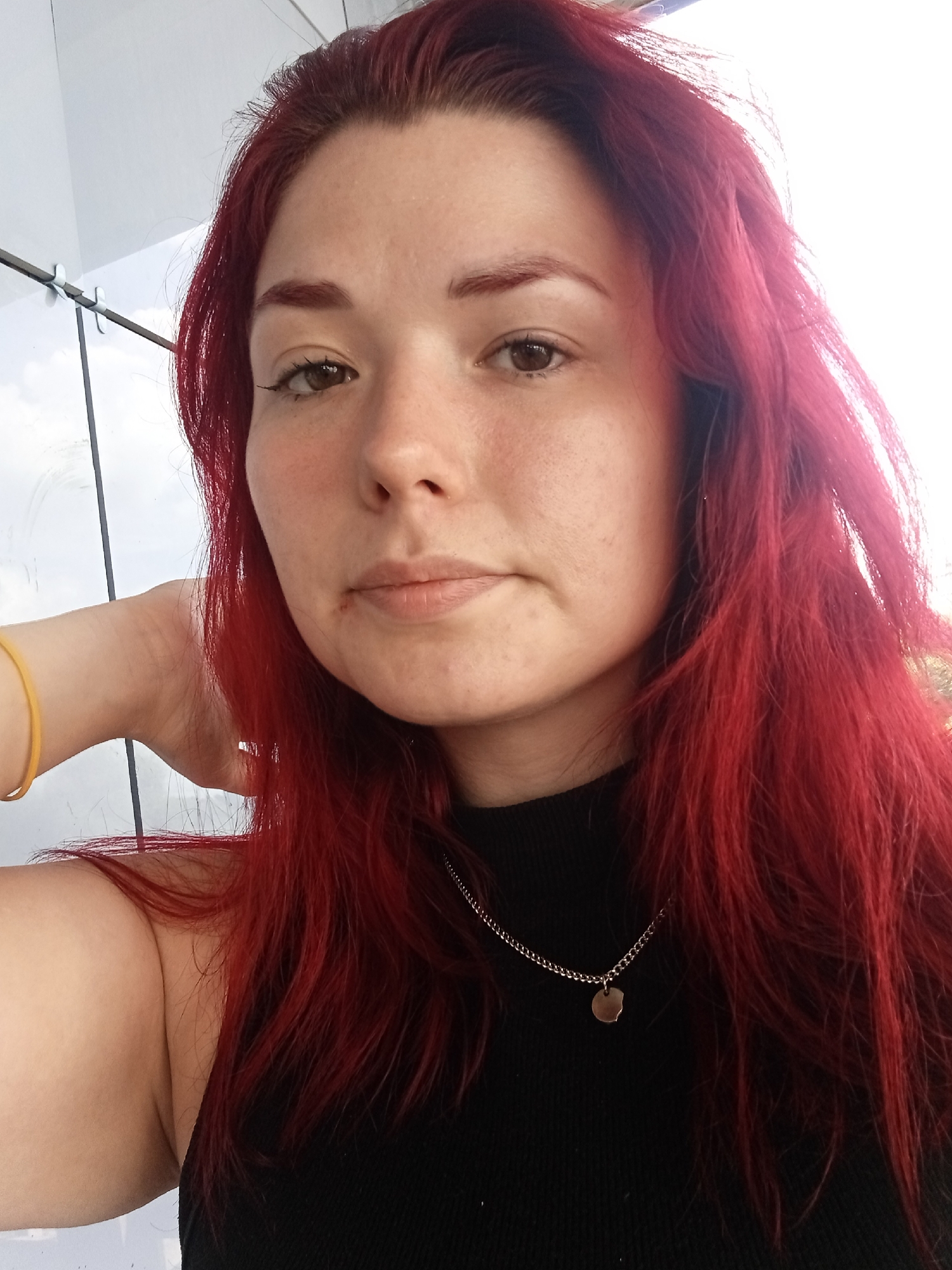 A medium shot portrait of a young woman with vibrant red hair is captured against a bright, overcast sky and a reflective surface, possibly a glass wall or building. She is looking directly at the camera, her right hand resting on the back of her neck. Her skin is fair with some freckles and blemishes visible. She has light brown eyes and dark red eyebrows that are slightly darker than her hair. She is wearing a black, high-necked garment and a thin, gold chain necklace with a small, round pendant. A yellow bracelet is visible on her left wrist. The background suggests an urban outdoor setting, though the specific location in Chaikovsky, Russia cannot be determined from the visual information provided.