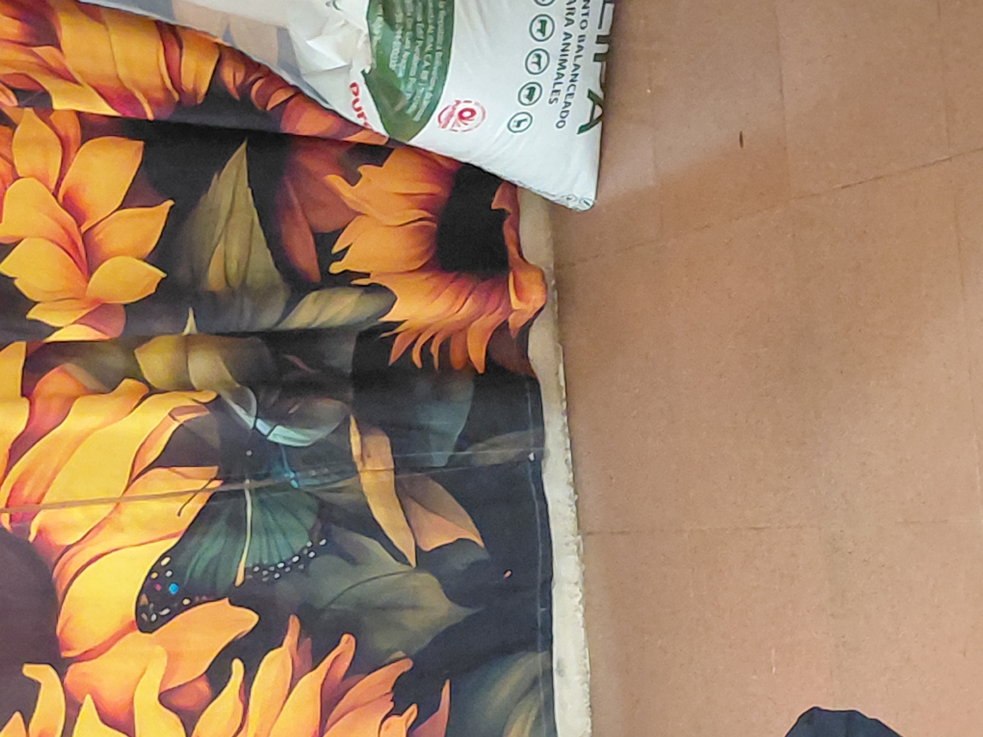 The image shows a close-up of a colorful tapestry featuring sunflowers and a butterfly. To the right of the tapestry is a white bag of animal feed with Spanish text. The bag is partially visible and has green and red lettering. The floor beneath the tapestry and bag appears to be tiled with a light brown material. There are no people visible in the image. The scene appears to be indoors. Without additional context, the exact location in Santa Ana, Venezuela, cannot be determined.