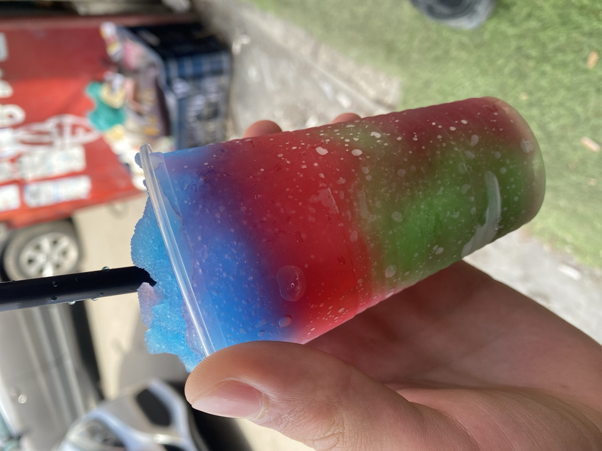 A person is holding a clear plastic cup filled with a layered frozen dessert in their hand. The dessert is colored blue at the bottom, red in the middle, and green at the top. A black straw protrudes from the blue layer. The cup has condensation on the outside. The person's thumb is visible holding the cup.
In the background, partially obscured, is a vehicle, possibly a car or truck, with a white and silver color scheme. Further in the background is a patch of green grass. The setting appears to be outdoors. The location within the city of Varna, Bulgaria cannot be determined from the context.