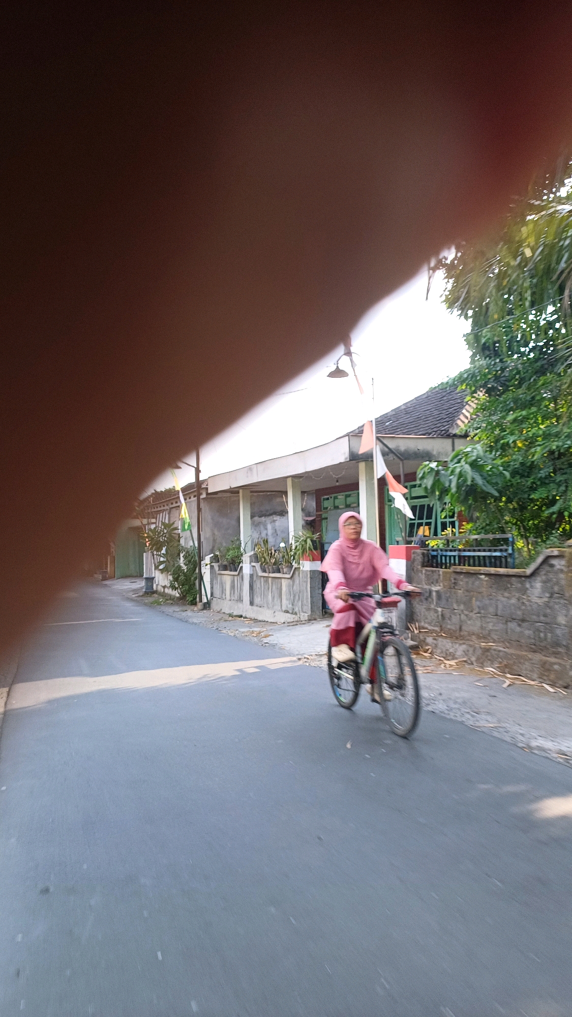 In Gergunung, Indonesia, on a paved street, a person wearing a pink hijab and matching outfit rides a bicycle. The person is blurred due to motion, but their form and the bicycle are discernible. The street is bordered by a low stone wall on the right and a row of buildings with white awnings on the left. Trees and foliage are visible behind the wall and to the right of the buildings. Two Indonesian flags are mounted on poles in front of the buildings. The lighting suggests it is daytime, with sunlight casting shadows on the street. A large, out-of-focus, brown object, likely a hand or finger, partially obscures the upper left portion of the image.
