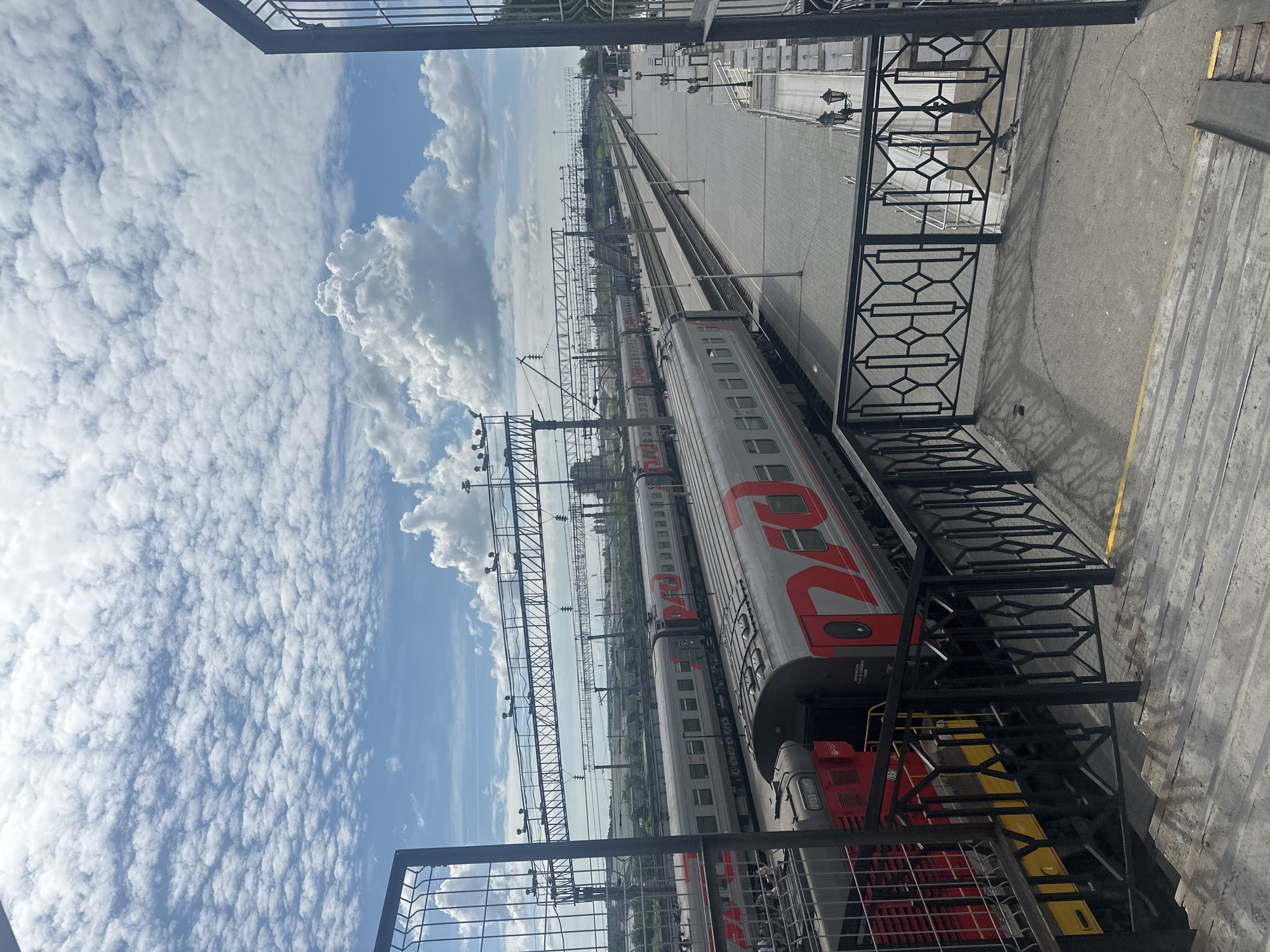 The image depicts a panoramic view of the Izhevsk railway station in Russia, observed from an elevated platform or pedestrian overpass during daylight hours under a sky with both widespread stratocumulus clouds and larger cumulus formations.
In the immediate foreground, a concrete staircase descends towards a lower level, bordered by a black metal railing with repeating geometric patterns. A yellow line marks the edge of the walking surface.
The midground is dominated by multiple parallel train tracks and station platforms. On the track closest to the viewer, a gray passenger train with a prominent red "РЖД" (RZD) logo is stationary. Text on its side reads "ПРИПИСНОЙ ПУНКТ УЧЕТА ИЖЕВСК ГОРЬК 17400," directly confirming "Izhevsk." Partially visible to the left of this train is the front section of a red and yellow ЧМЭ3-series shunting locomotive, bearing the number "ЧМЭ3-1057." Further down the tracks, more gray passenger cars with the red RZD logo are visible.
Several individuals are present on the paved platforms between the tracks, appearing small due to the viewing distance. Overhead, a complex network of electrical catenary lines and support gantries spans across all tracks, indicative of an electrified railway system. Beyond the immediate tracks, a pedestrian overpass traverses multiple lines.
In the distant background, industrial or urban structures are visible, and beyond them, a broad, flat expanse of what appears to be a large body of water, likely the Izhevsk Pond, stretches to the horizon. Lampposts with multiple light fixtures are regularly spaced along the platforms. The overall scene conveys a functional, active railway environment.