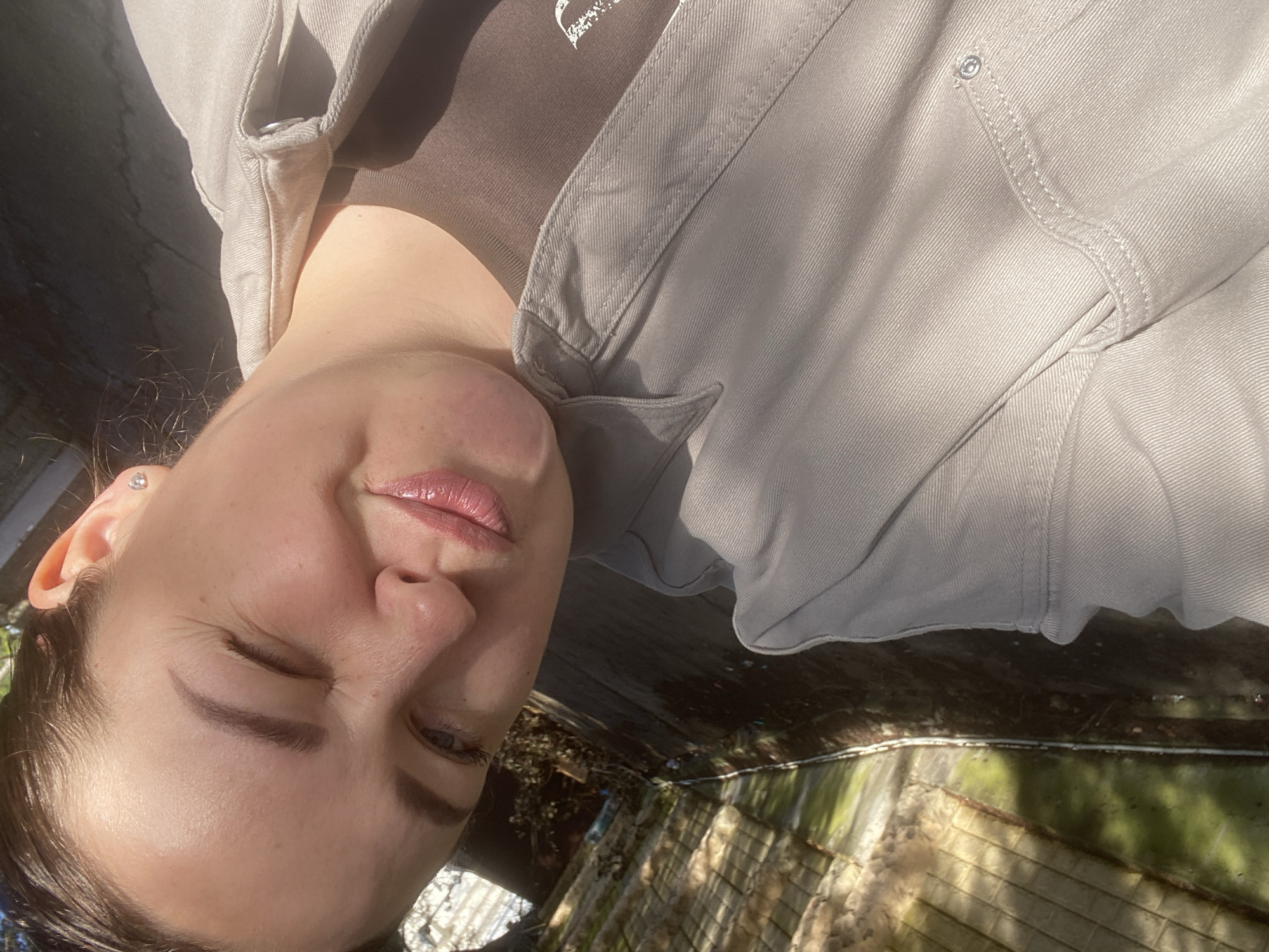 A close-up, tilted portrait captures a young woman outdoors in Sochi, Russia, on a bright, sunny day. The subject is positioned with her head to the left, looking towards the camera with her eyes mostly closed or squinting, likely due to the intense sunlight. Her lips are slightly parted and enhanced with glossy pink lipstick. A small, clear stud earring is visible in her left ear.
She is wearing a light grey or beige jacket, possibly denim or a similar sturdy fabric, with visible stitching details, a collar, and a metal button on a chest pocket. Underneath, she wears a dark brown top, which features a small, stylized white design, resembling a heart.
The background is out of focus, indicating an outdoor setting with strong highlights and shadows. Reflections of sunlight are prominent on what appears to be a water feature or pond, with hints of green vegetation. The overall impression is one of leisure, with the woman seemingly enjoying the warmth and brightness of the day.