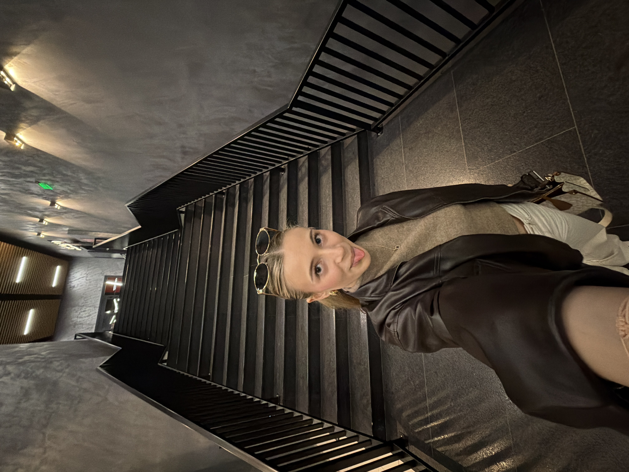 A young Caucasian female is depicted taking a selfie on a modern indoor staircase within a building in Plovdiv, Bulgaria. She is positioned at the base of a flight of dark gray steps, which feature black metal railings with vertical balusters on both sides. The walls adjacent to the staircase are dark gray and textured. The subject wears a dark brown leather jacket over a light brown turtleneck or high-neck sweater, and white trousers. Gold-rimmed oval sunglasses rest on top of her head, and she wears gold hoop earrings and a delicate gold necklace. Her mouth is open with her tongue extended. Her left hand holds a light-colored patterned shoulder bag. The environment is illuminated by several rectangular wall-mounted sconces on the right wall, casting light upwards and downwards, and an illuminated green exit sign is visible above them. At the top of the staircase, a wooden slatted ceiling with rectangular recessed lights is visible, along with a doorway featuring a prominent red cross-shaped light. The floor at the subject's feet is composed of dark gray tiles.