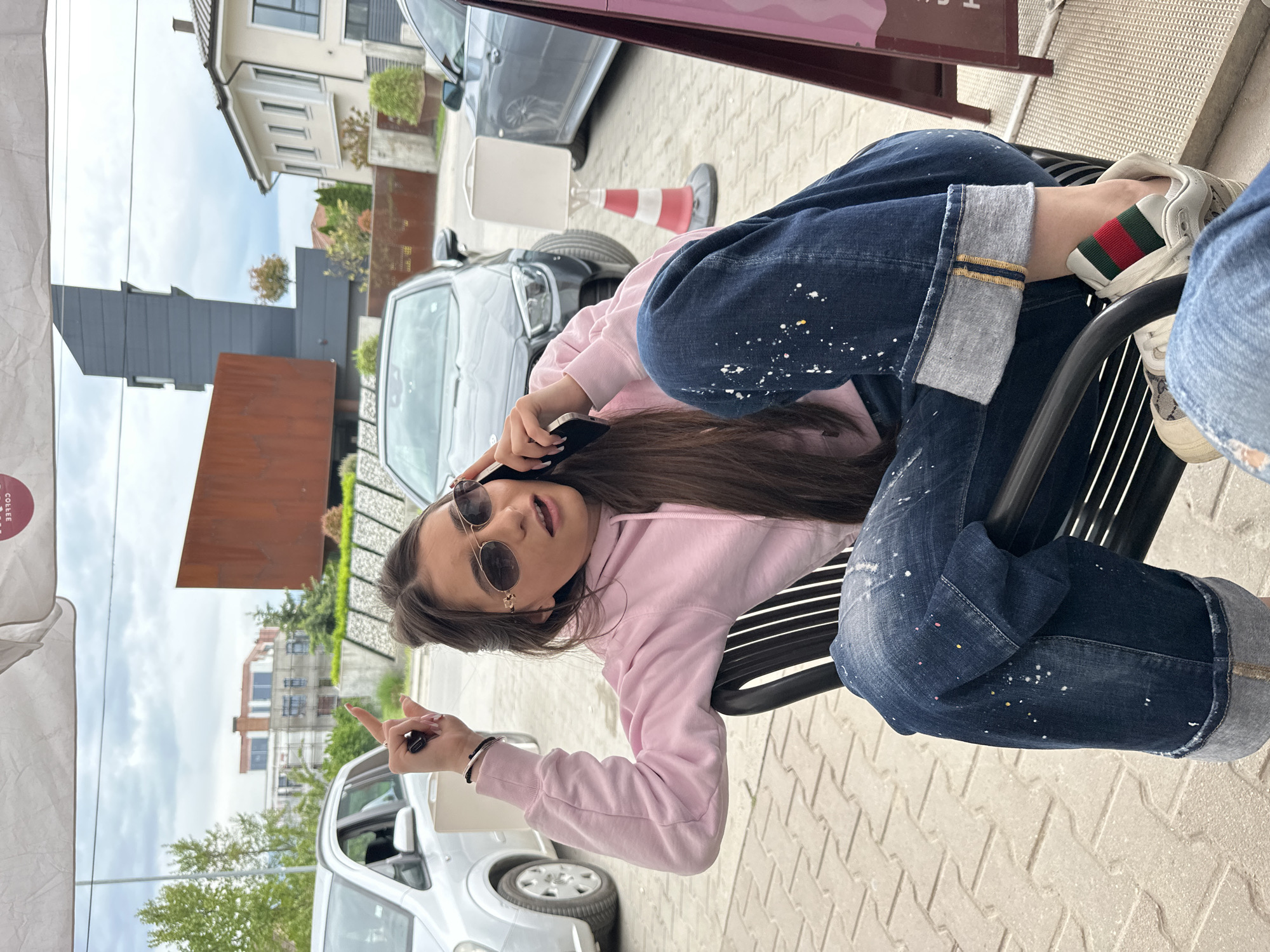 A young woman is reclining on a black slatted outdoor bench. She wears sunglasses, a pink hoodie, and splatter-painted blue jeans cuffed to reveal a tan fabric underneath, and Gucci sneakers. She is holding a smartphone to her ear and gesturing with her left hand. Cars and buildings are visible in the background. The location may be Plovdiv, Bulgaria, but the specific location is not identifiable from the image. The ground is paved with light-colored bricks.