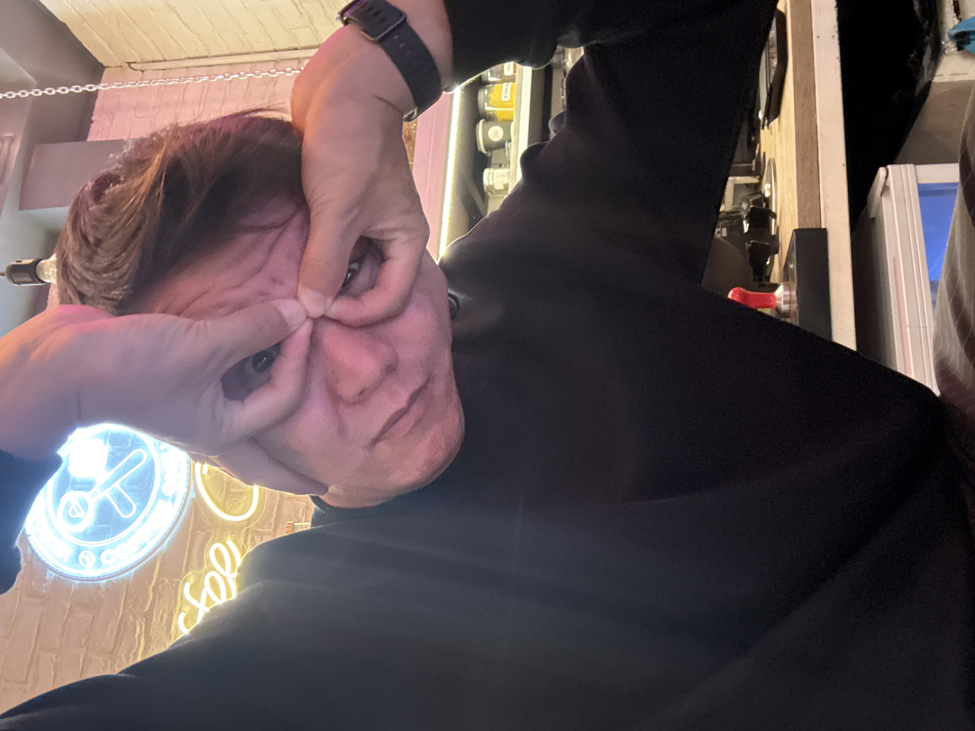 In Moscow, Russia, a person is captured in a close-up shot, their face contorted as they pull their skin near their eyes. They are wearing a black long-sleeved shirt and a black wristwatch on their right wrist. The background features a brick wall with neon signs. One sign, illuminated in blue, displays a circular design with an object resembling a whisk. Another neon sign, in yellow, spells out "coffee". Shelves in the background hold various containers, some appearing to contain yellow powder. A light fixture is visible above. The overall scene suggests an indoor setting, possibly a cafe or bar.