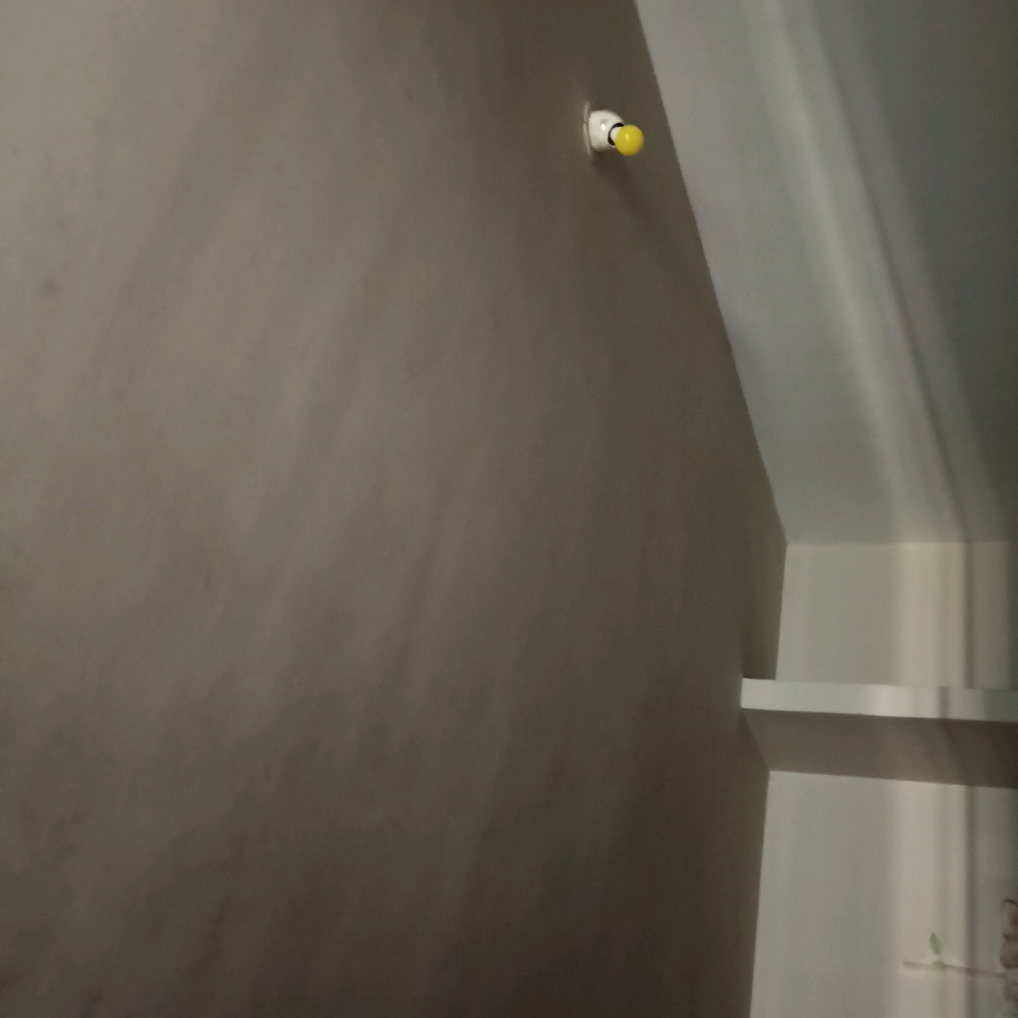 The image depicts an interior wall with a light fixture. A small, yellow bulb is visible in a white casing attached to the wall. The wall appears to be painted a neutral, gray tone. To the right of the wall, a portion of a white shelf is visible, suggesting the presence of shelving units. The lighting in the scene is dim, with some light reflecting off the white surfaces. There are no people or animals visible in the image, nor any objects that would indicate a specific location or activity. The scene is devoid of any discernible elements that would place it in Bengaluru, India, or any other specific city.