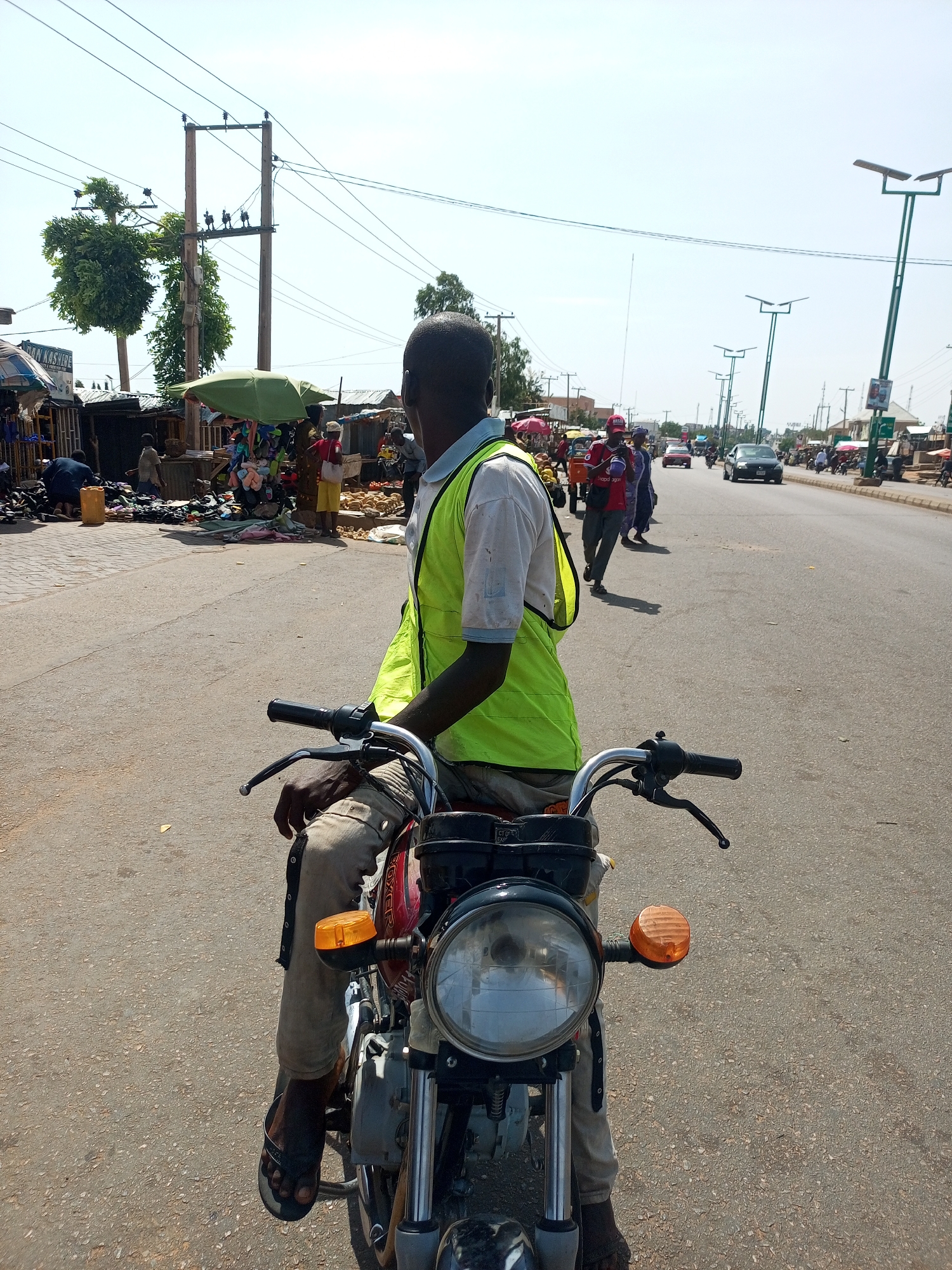A man on a motorcycle, viewed from behind, is stopped or waiting on a paved road under a bright, clear sky. He wears a white short-sleeved shirt and a neon green reflective vest. His dark-skinned hands rest on the handlebars, and his feet, clad in sandals, are on the ground near the motorcycle. The motorcycle features a round headlight and prominent orange turn signals. The word "BOXER" is faintly visible on the fuel tank.
To the left of the road, a bustling market area is active with numerous people. Various stalls with corrugated metal roofs are visible, displaying merchandise such as shoes and what appears to be produce like coconuts or yams. Green umbrellas provide shade over some of the market stands. Overhead, several utility poles with a dense network of power lines stretch across the scene.
To the right, the paved road extends into the distance, with several cars and motorcycles visible in traffic. Modern streetlights, possibly solar-powered, line the side of the road. The strong sunlight casts distinct shadows, indicating it is likely midday. The scene depicts a lively, everyday street and market environment in what appears to be a populated area, possibly in an African setting. Text "DAN KASHERE" is visible on one of the market stall umbrellas.