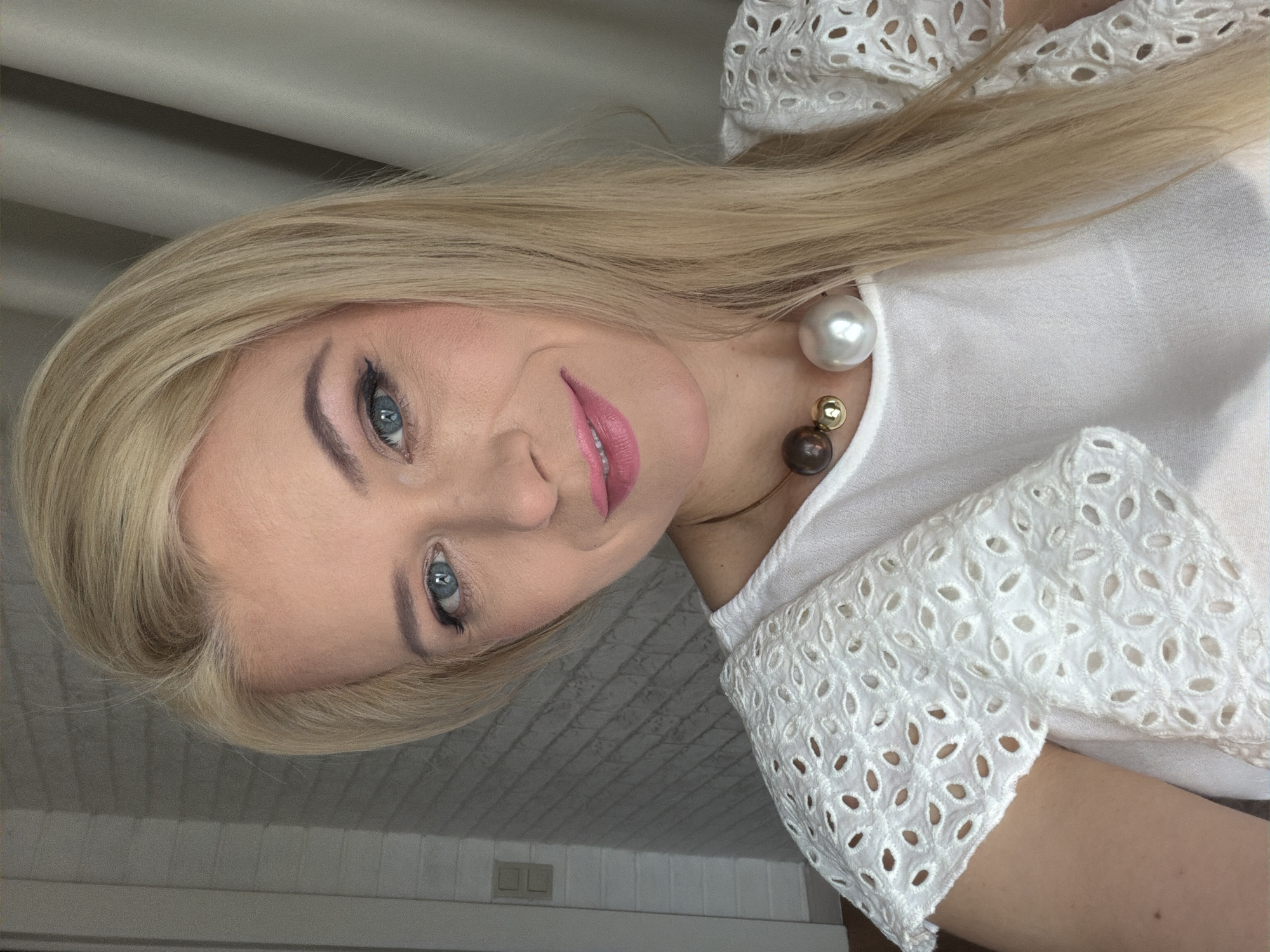 A close-up, waist-up, indoor, selfie-style portrait of a Caucasian woman with blonde hair and blue eyes. She is wearing a white, eyelet-lace top with ruffled sleeves and a multi-colored beaded necklace. The woman is smiling and looking directly at the camera. The background features a light-colored brick wall and a beige curtain. The image appears to have been taken in Sochi, Russia.