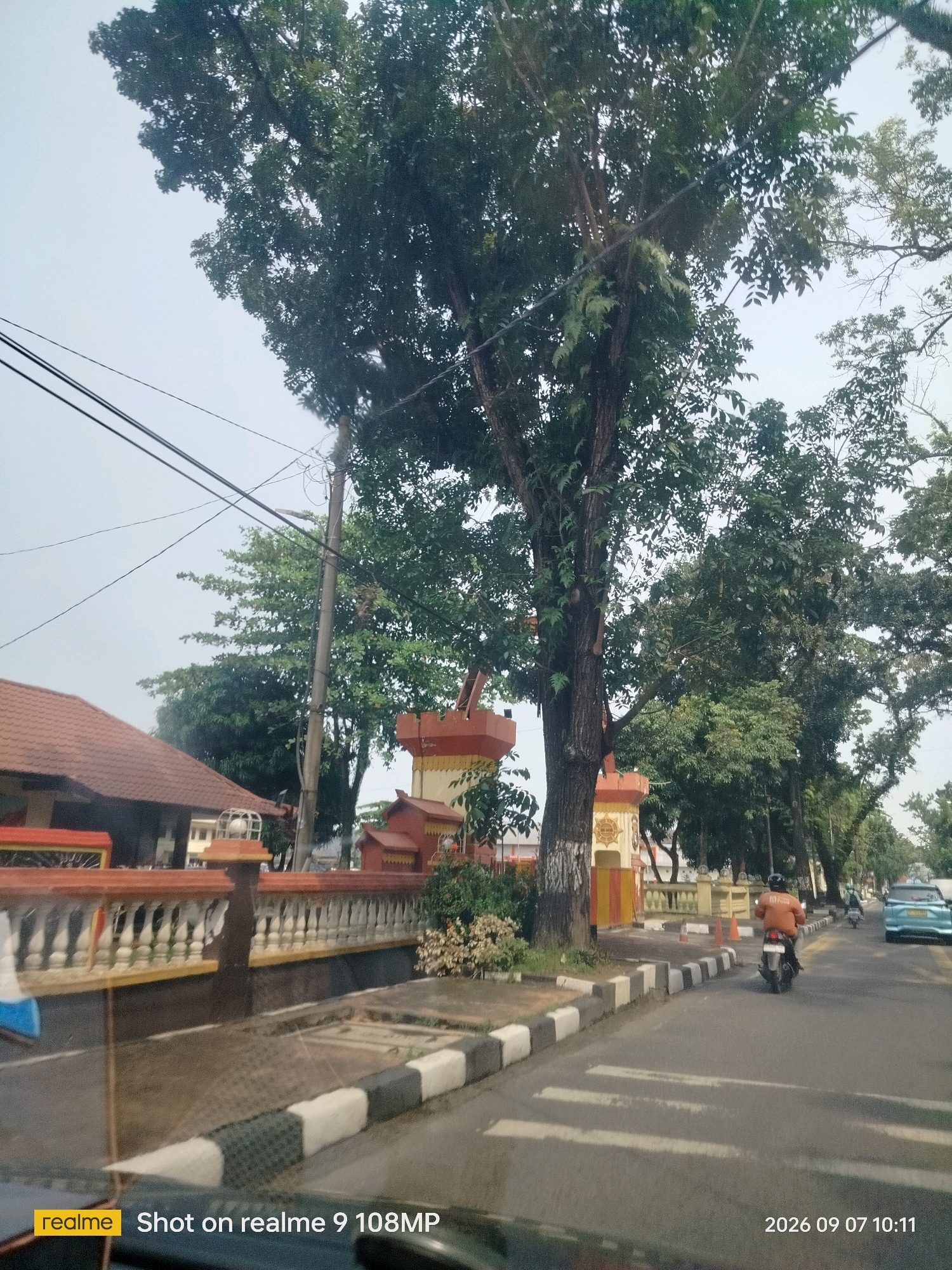 The image captures a street scene in the city of Medan, Indonesia, on September 7, 2026, at 10:11 AM. A large tree with dense green foliage dominates the foreground on the left, casting shadows on a striped pedestrian crossing. A white and black striped curb lines the street, separating it from a sidewalk.
On the sidewalk, a motorcycle rider wearing an orange jacket is seen from behind, riding away from the camera. To the right of the motorcycle, a light blue car is visible in the distance. Beyond the sidewalk, a building with a reddish-brown tiled roof is partially visible, featuring a decorative fence with white balusters. Further down the street, several other trees and a utility pole with overhead wires are present. The sky is overcast.
The timestamp "2026 09 07 10:11" and the text "realme Shot on realme 9 108MP" are overlaid at the bottom of the image.