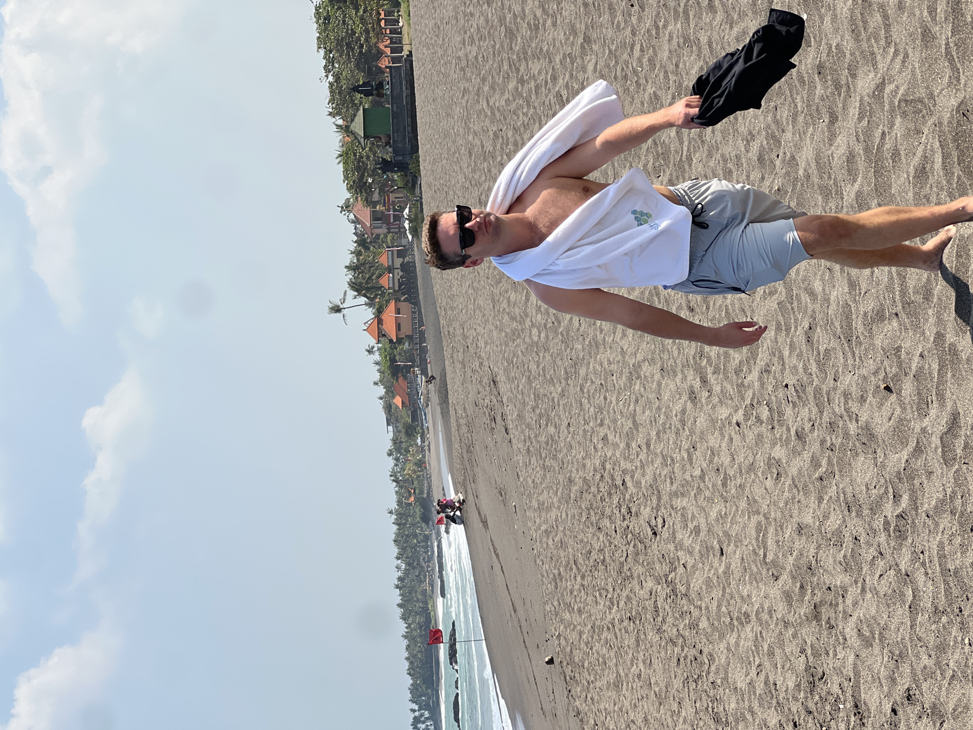In Mengwi, Indonesia, a man with fair skin, blonde hair, and sunglasses walks on a dark sand beach. He is shirtless, wearing light grey swim trunks, and has a white towel draped over his shoulders. In his right hand, he carries a black t-shirt. The beach is mostly empty, with the ocean visible in the background, featuring small waves breaking on the shore. A few red flags are planted in the sand near the water's edge. In the distance, a line of palm trees and buildings with red roofs suggest a resort or village. The sky is light blue with scattered white clouds.